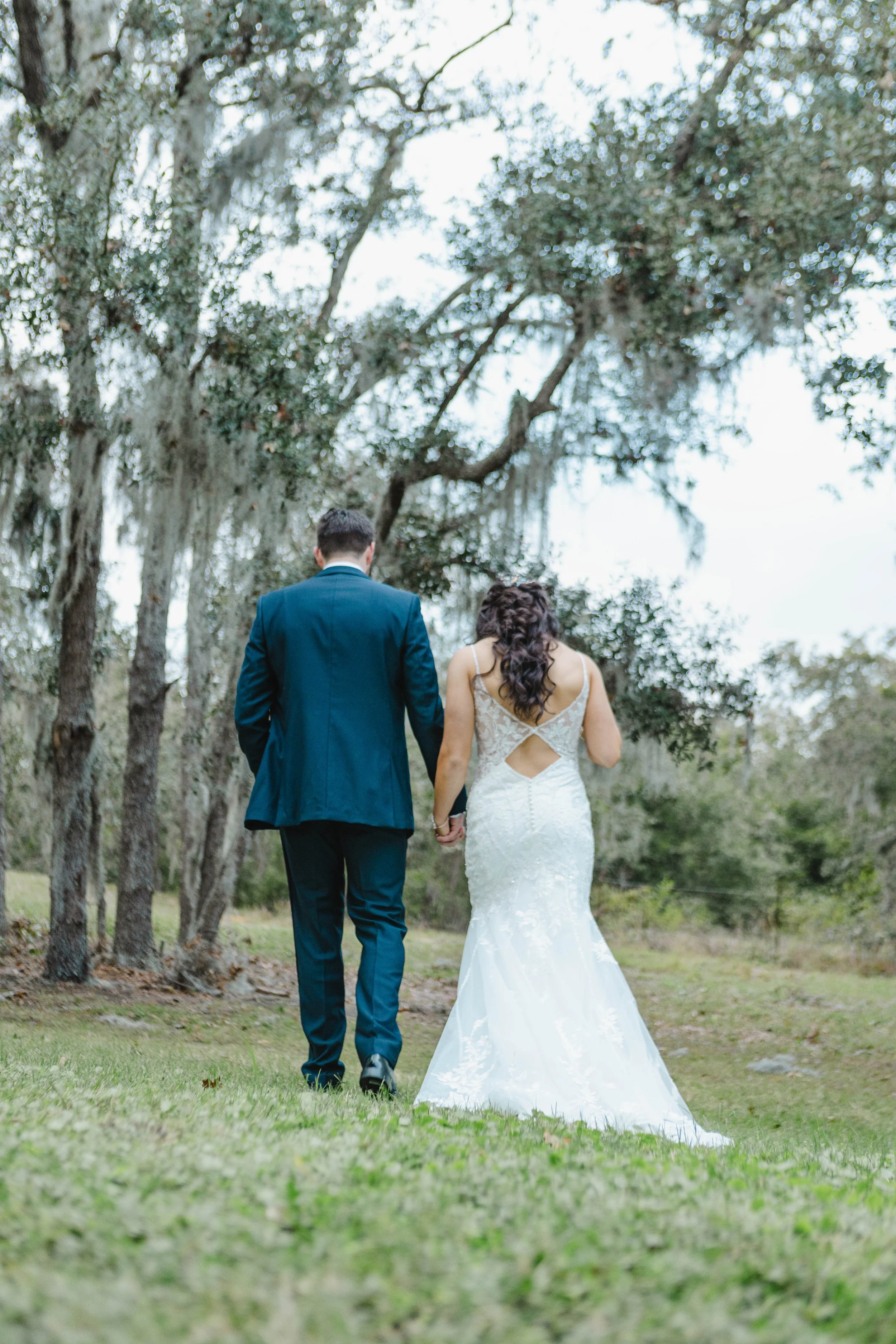 A bride and groom walking hand in hand outdoors among trees, as seen from behind. The bride is wearing a white lace wedding gown with a cutout back, and the groom is dressed in a dark suit.