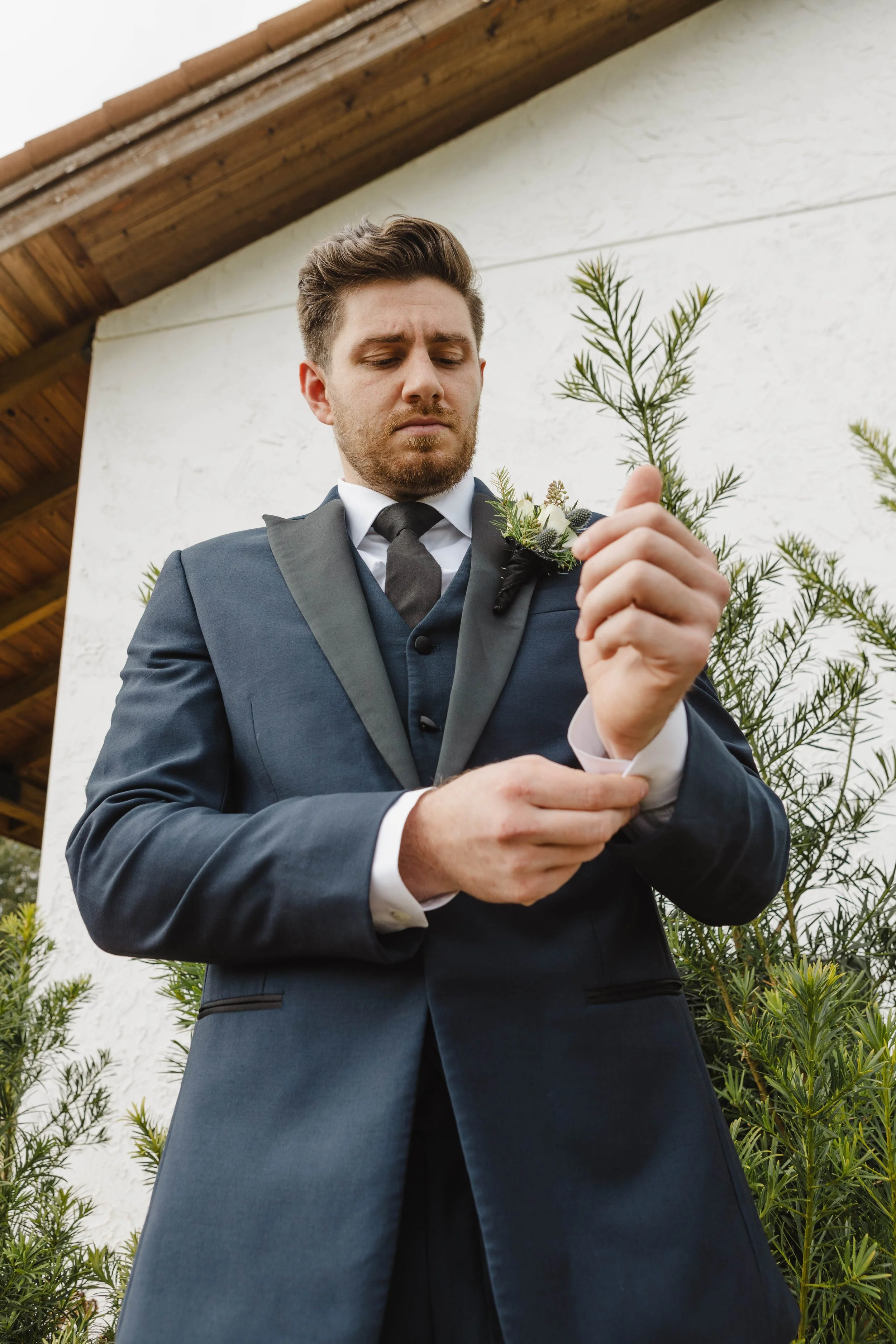 A man in formal attire adjusting his cufflink outdoors. He is wearing a dark suit with a boutonniere on his lapel, standing in front of some green bushes.