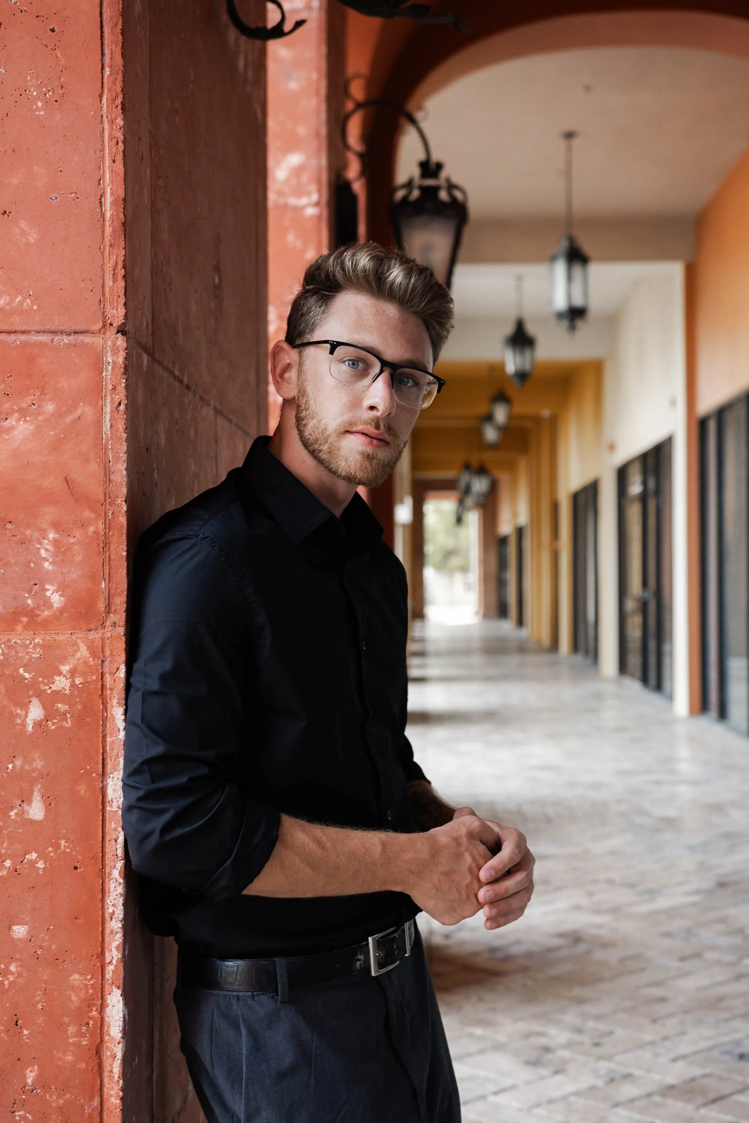A man with glasses and a beard leaning against a brick wall in a corridor with hanging lanterns and windows.