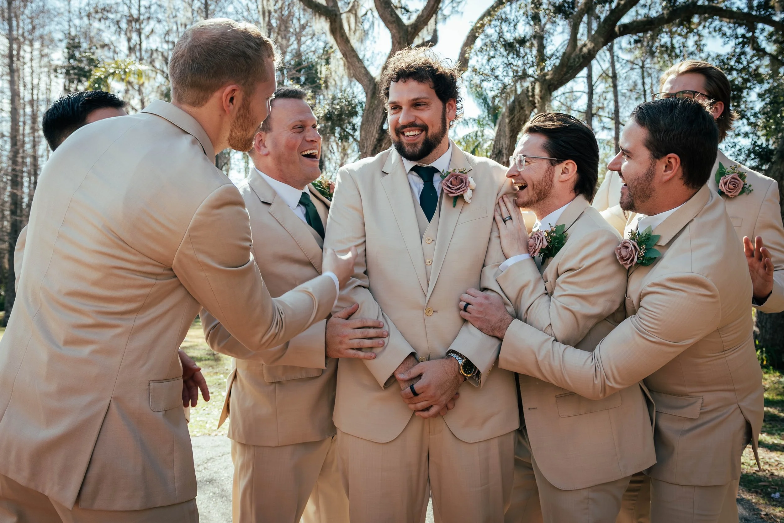 Group of men in beige suits smiling and laughing together outdoors with trees in the background, dressed for a wedding or formal event.