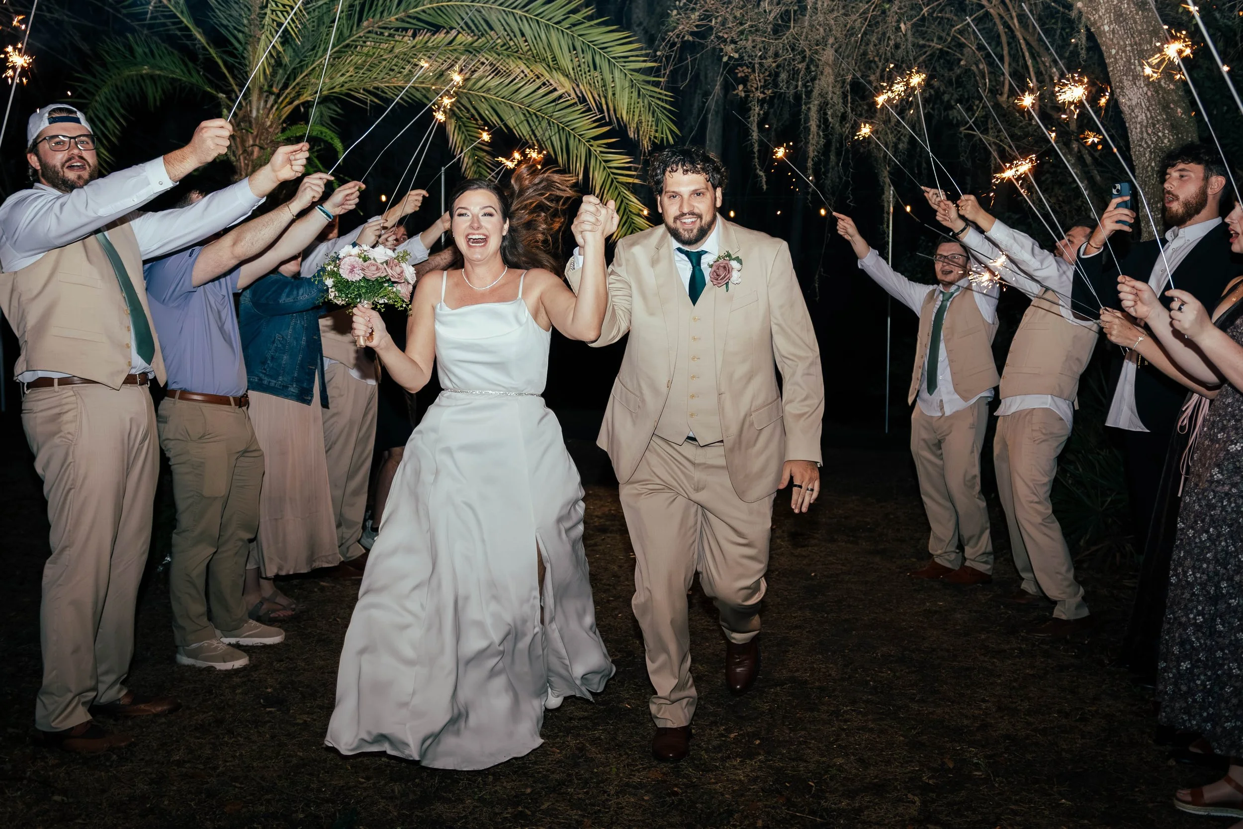 A couple in wedding attire walking under sparklers held by guests at night, surrounded by trees with hanging moss.