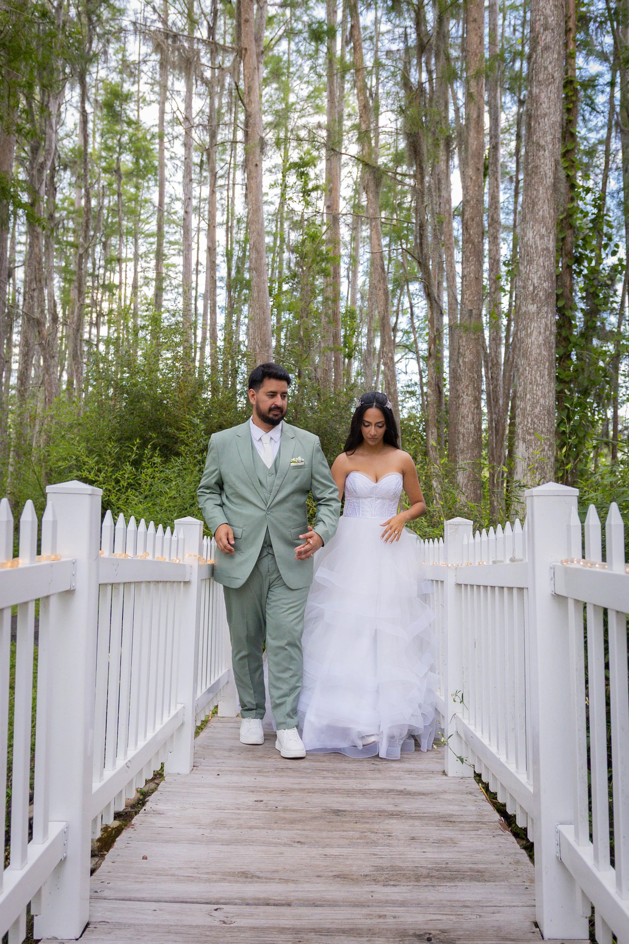 A bride and groom walking together across a white wooden bridge in a forested area during daytime.