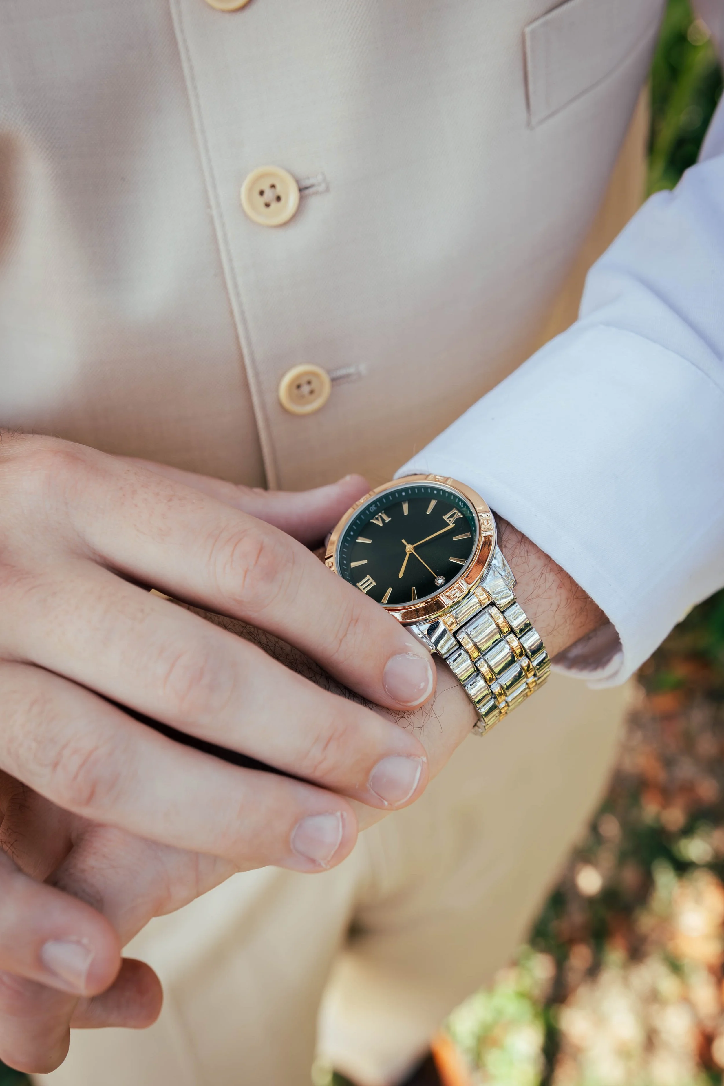Close-up of a person's wrist wearing a silver and gold wristwatch with a black face, with a beige vest with buttons and a white shirt sleeve visible.