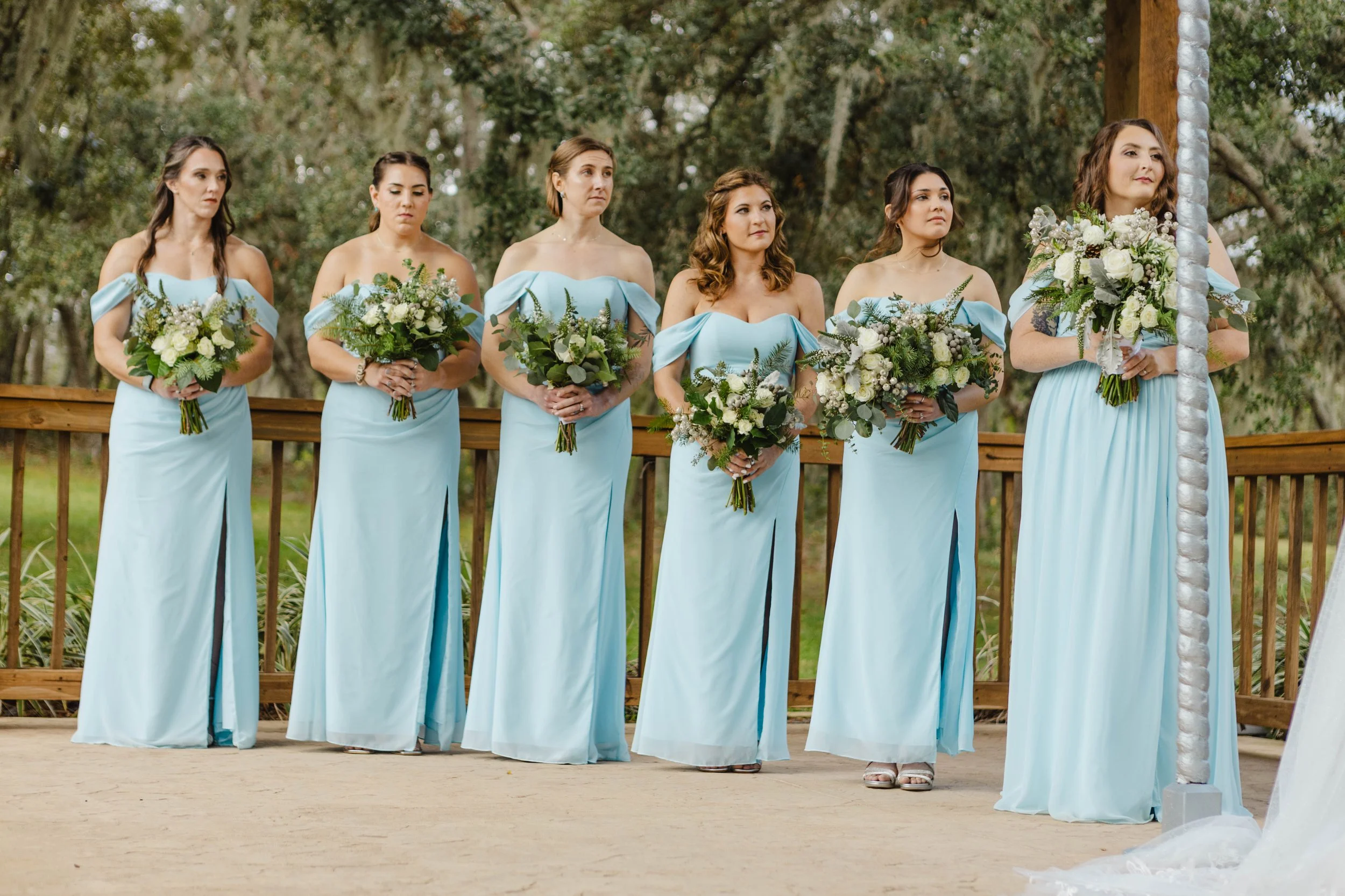 Six women dressed in light blue off-the-shoulder gowns holding bouquets of white flowers and greenery, standing outdoors on a wooden platform with trees in the background.