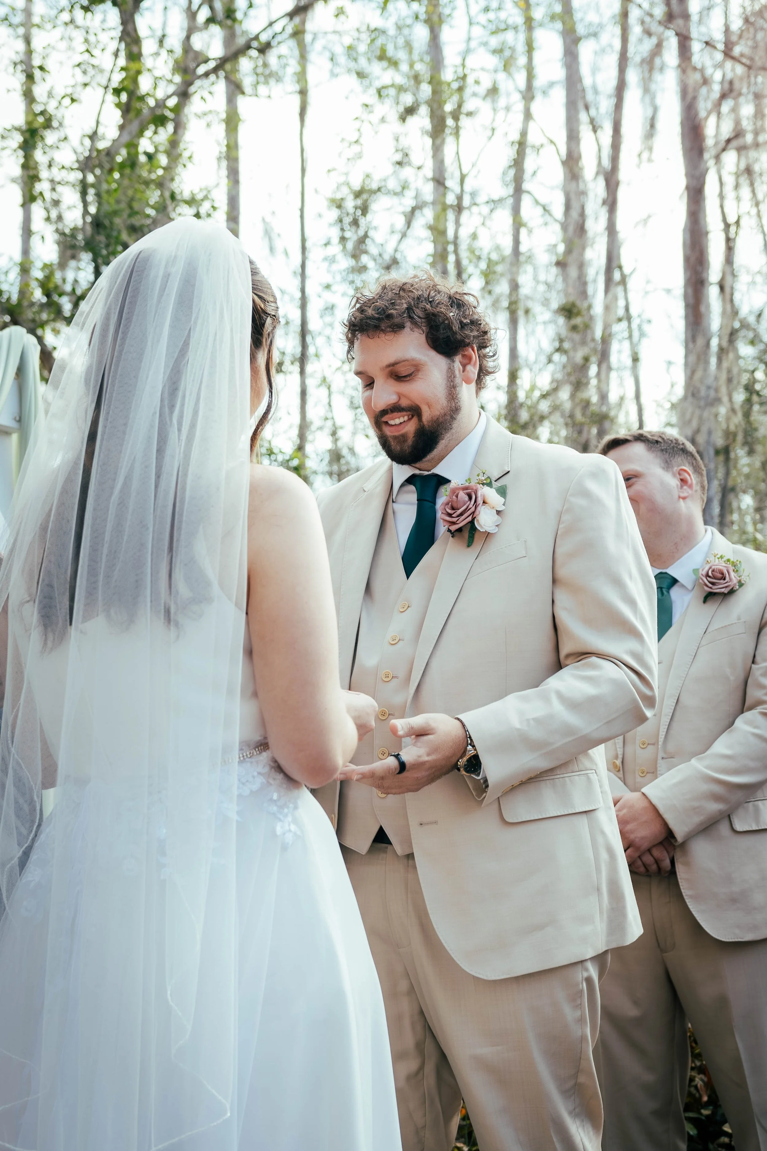 A wedding ceremony outdoors with a bride and groom exchanging vows, surrounded by green trees.