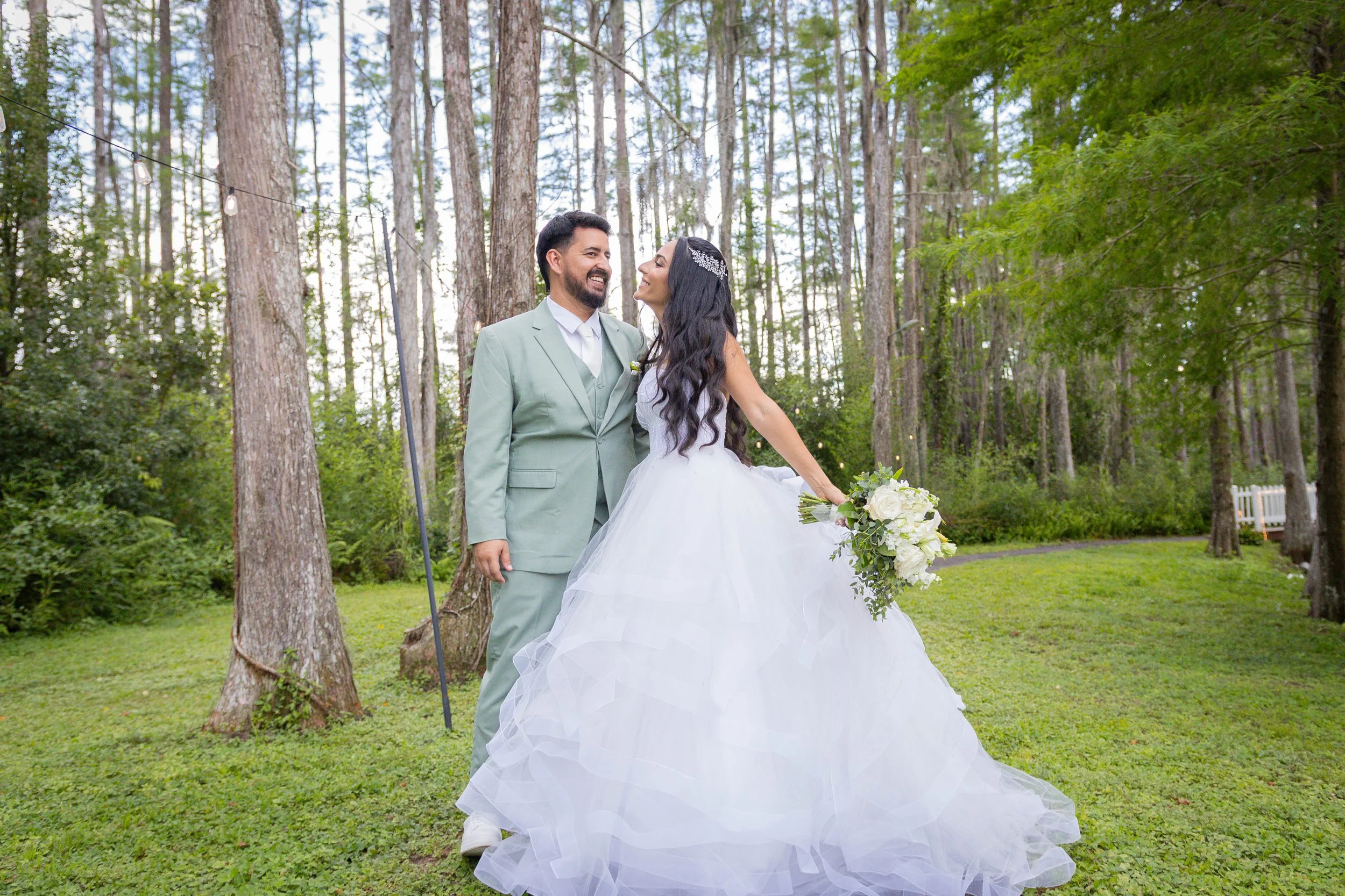 A smiling bride and groom in wedding attire standing in a wooded outdoor setting, facing each other with trees and string lights in the background.