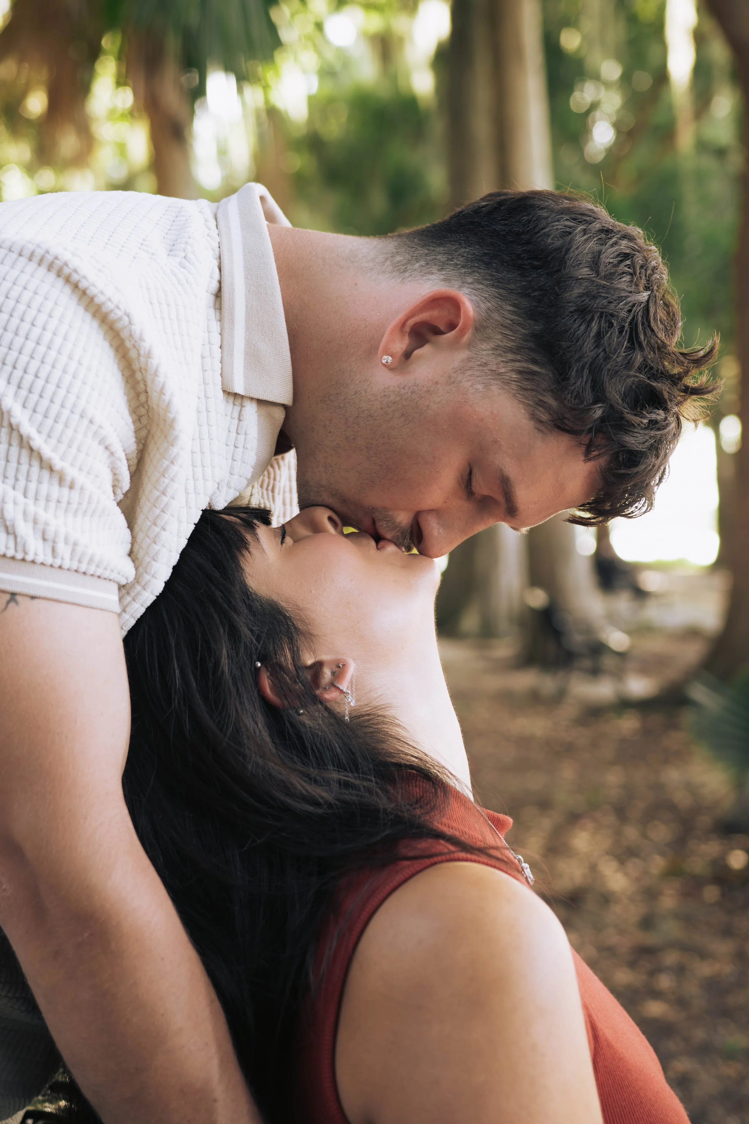 A man and woman sharing a kiss outdoors in a wooded area, with trees and sunlight in the background.