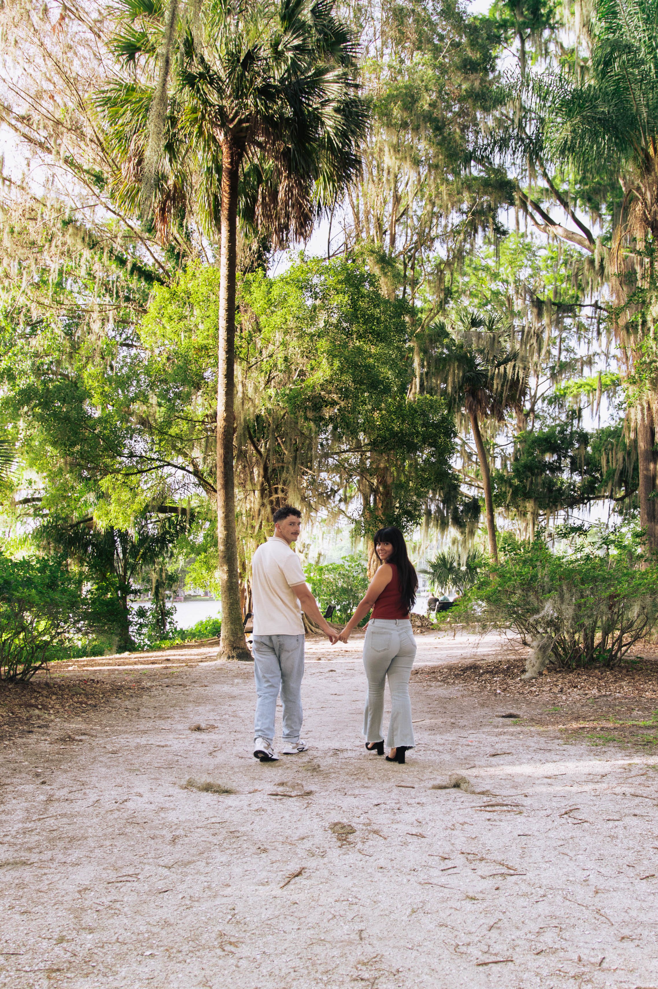 A young man and woman holding hands in a wooded park, surrounded by trees and greenery, with the water visible in the background.