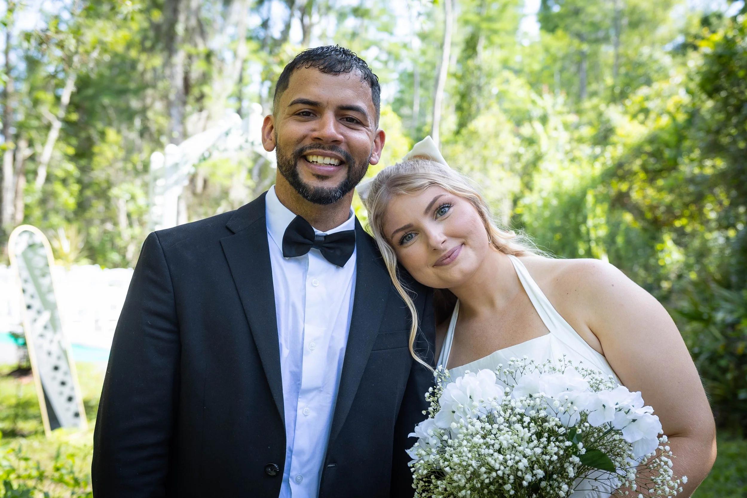A newlywed couple dressed in wedding attire, smiling and posing outdoors in a wooded area during daytime. The groom wears a black tuxedo with a bow tie, and the bride wears a white dress and holds a bouquet of white flowers.