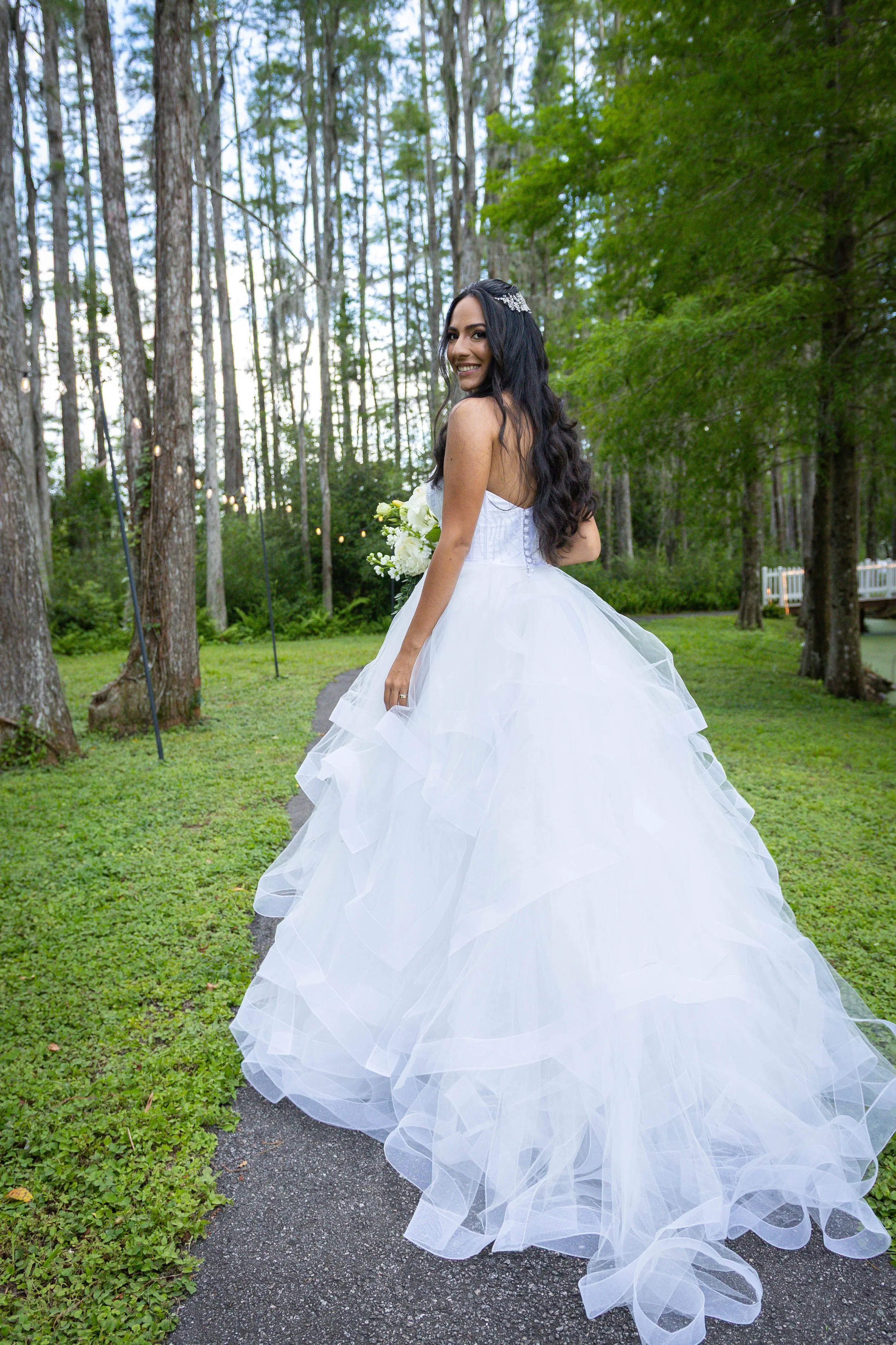 A woman in a white wedding dress with a long, flowing skirt is standing on a paved path in a wooded outdoor setting, holding a bouquet of white flowers and smiling at the camera.