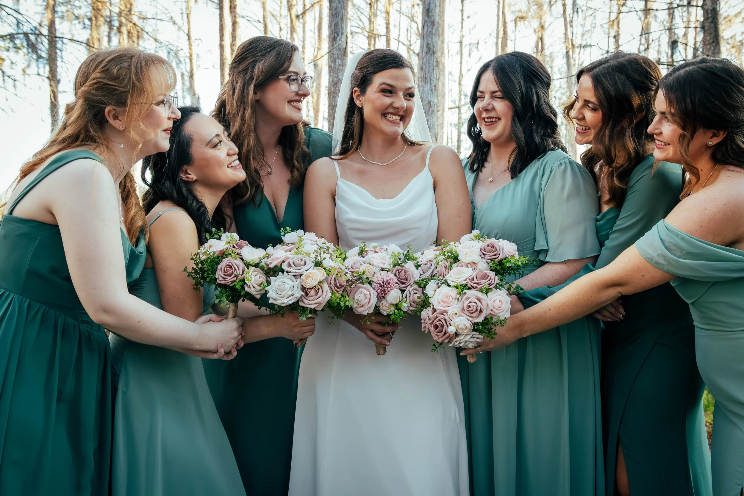 A bride in a white dress smiling surrounded by her bridesmaids in teal dresses, holding a bouquet of pink and white roses, outdoors in a forest setting.