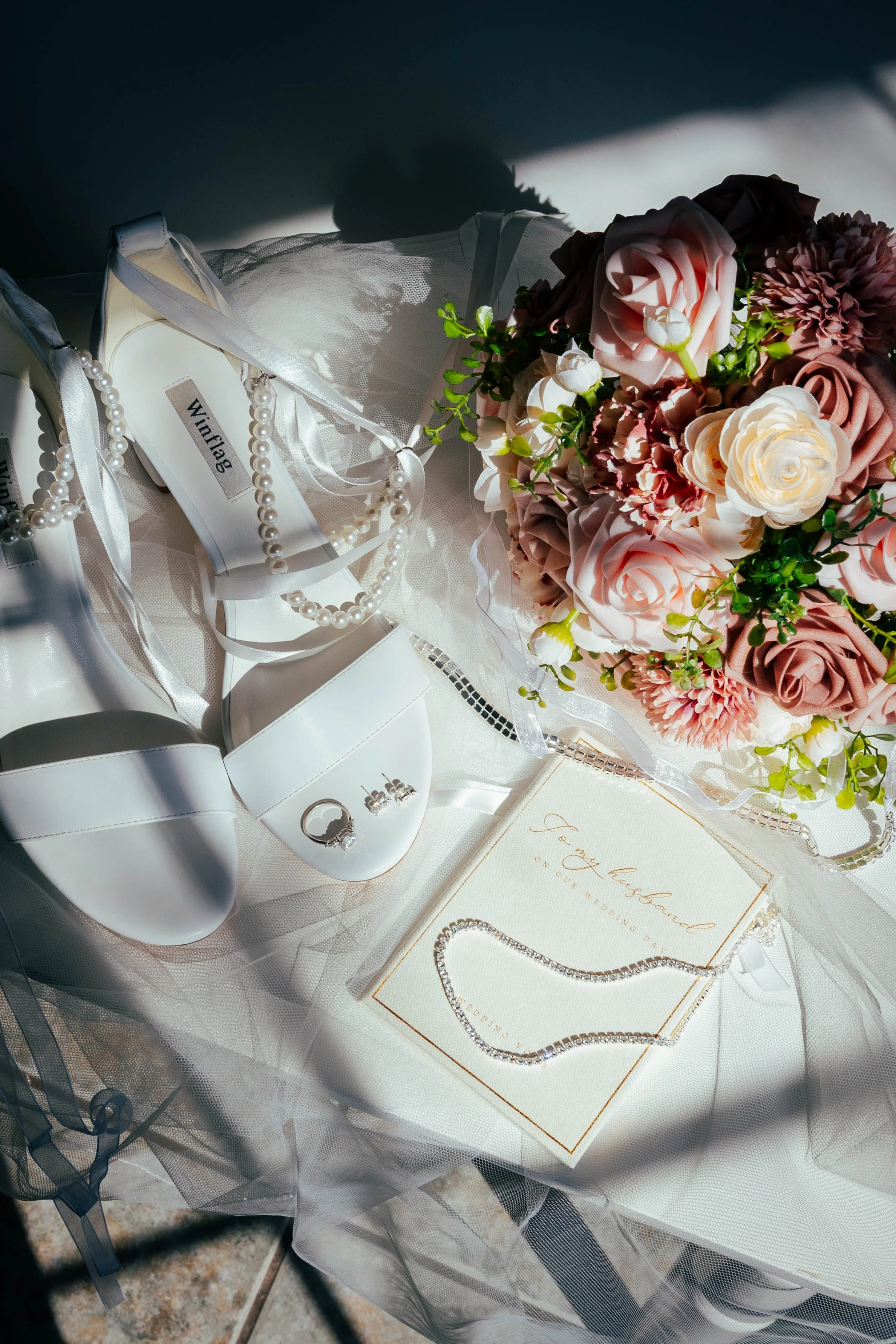 Wedding accessories including white high-heeled shoes with pearl straps, a pearl necklace, a silver ring, a white veiled guest book with gold lettering, a string of diamond jewelry, a bridal bouquet with pink, white, and mauve roses, and a wedding in