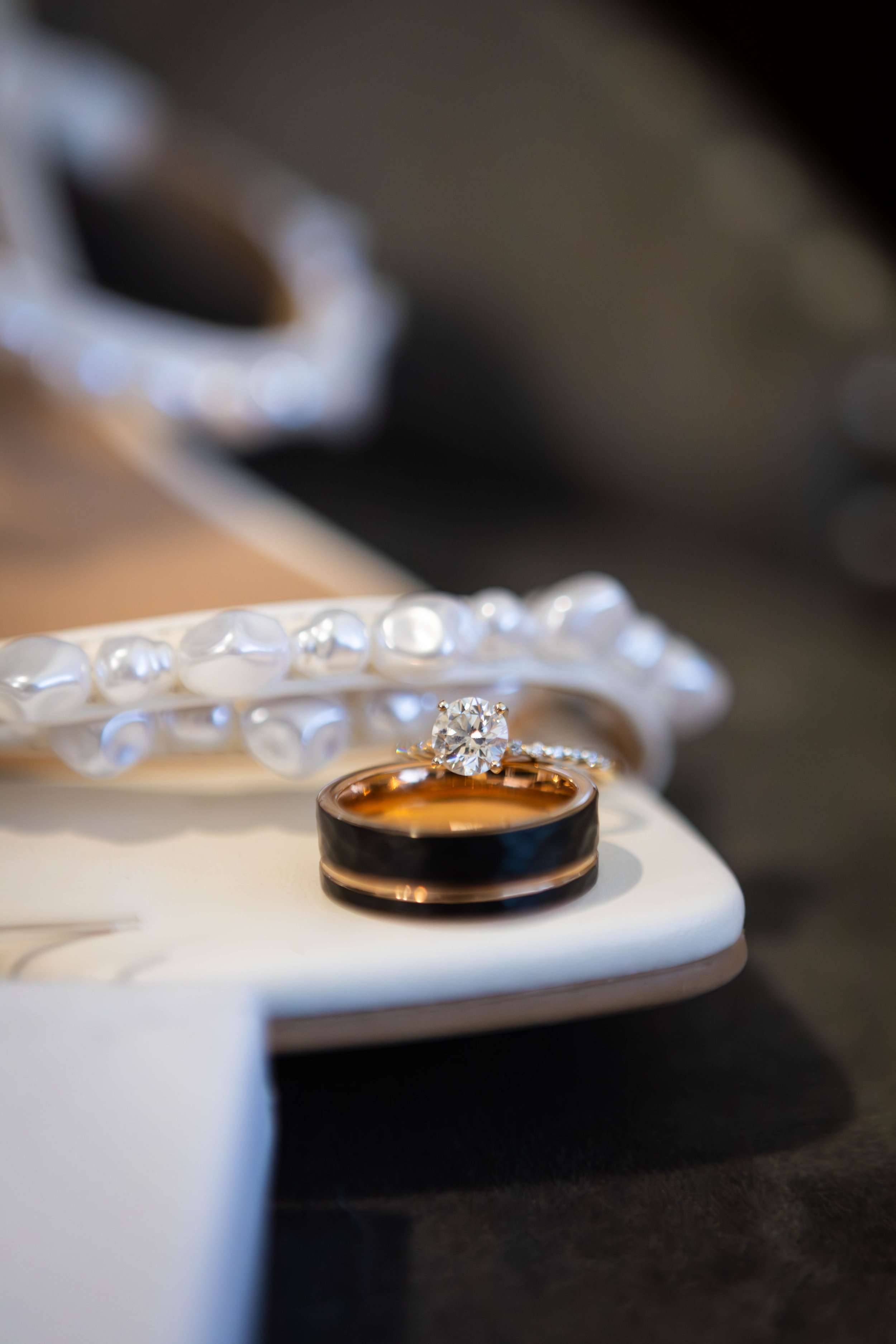 Close-up of a jewelry display with a diamond ring, a black and gold ring, and a pearl bracelet on a white stand.