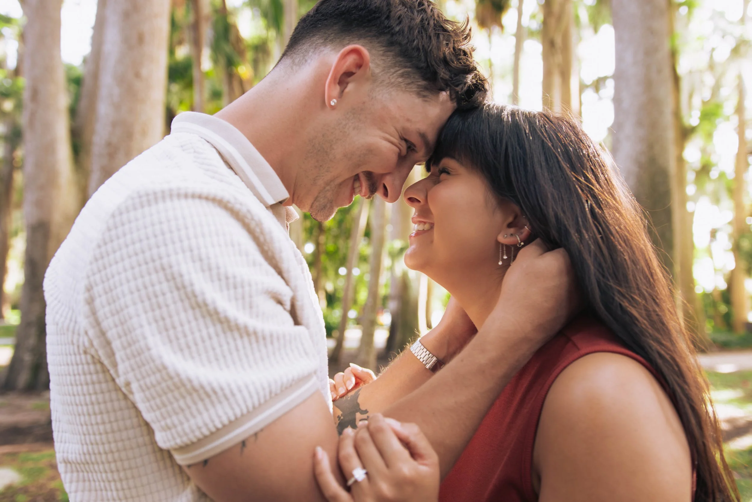 A man and woman are touching foreheads and smiling in a park with trees in the background.