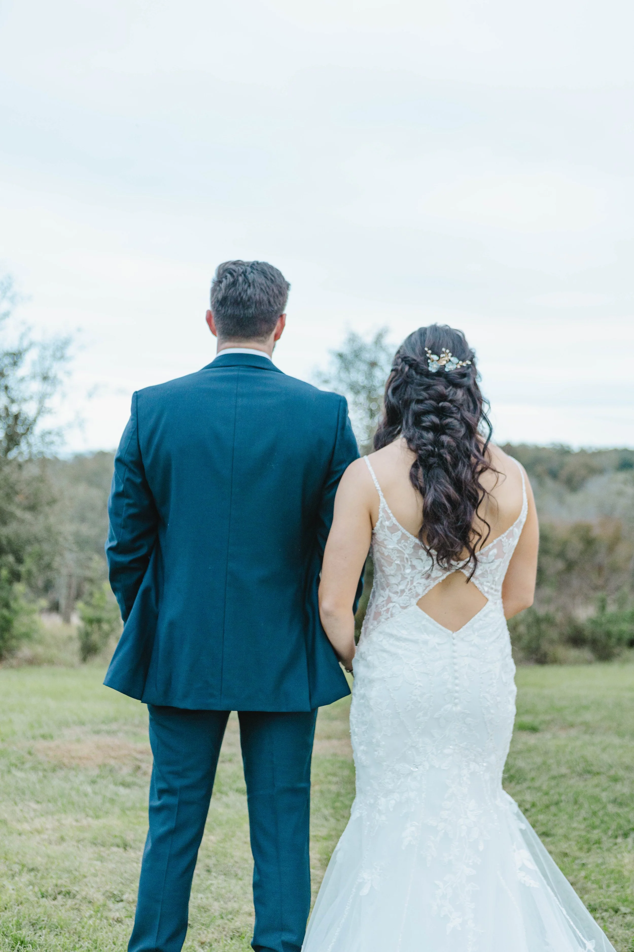 A bride and groom stand outdoors at a wedding, seen from behind, with the groom in a dark blue suit and the bride in a white lace wedding dress with a cutout back and floral hair accessories.
