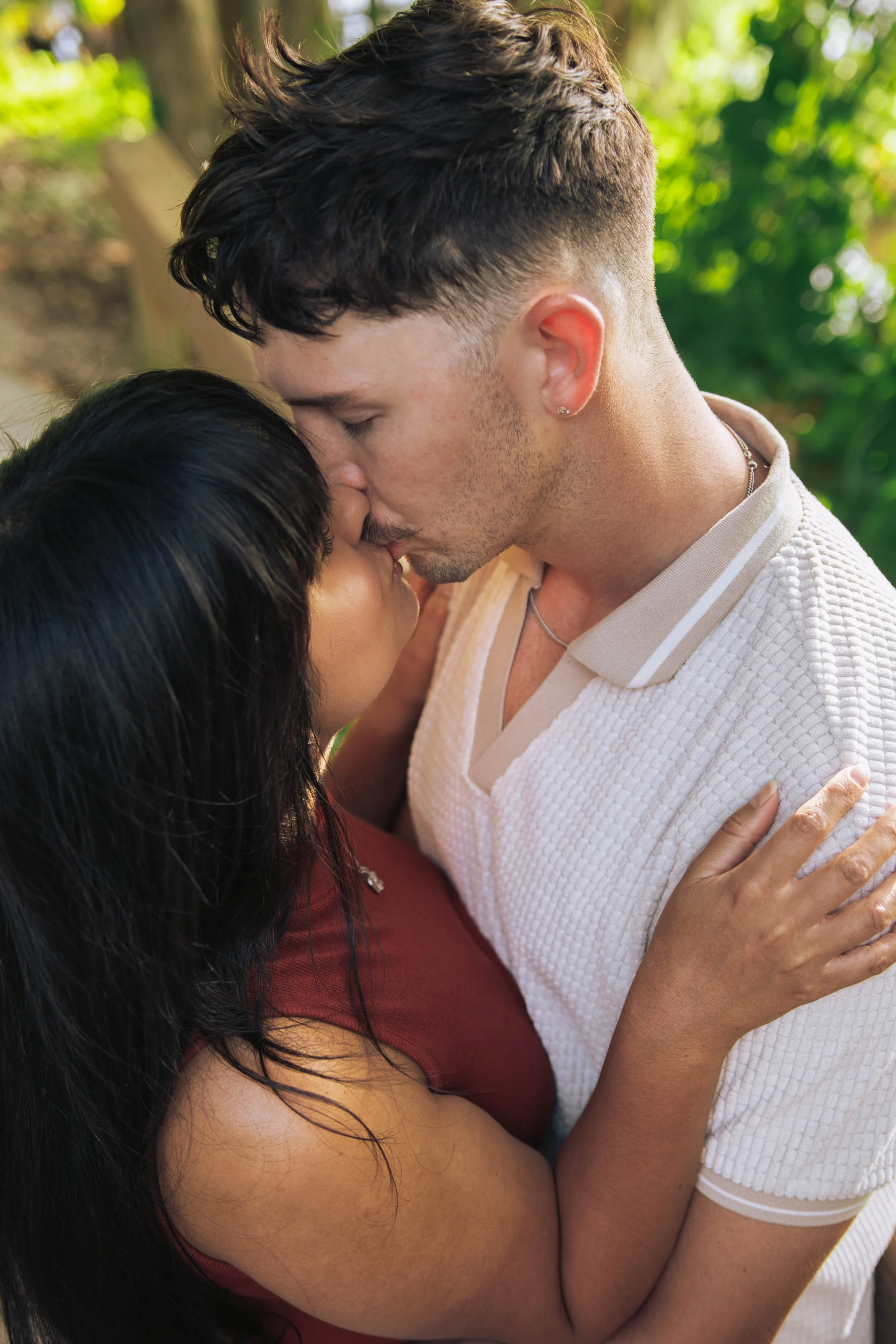 A man and woman are kissing outdoors with greenery in the background.