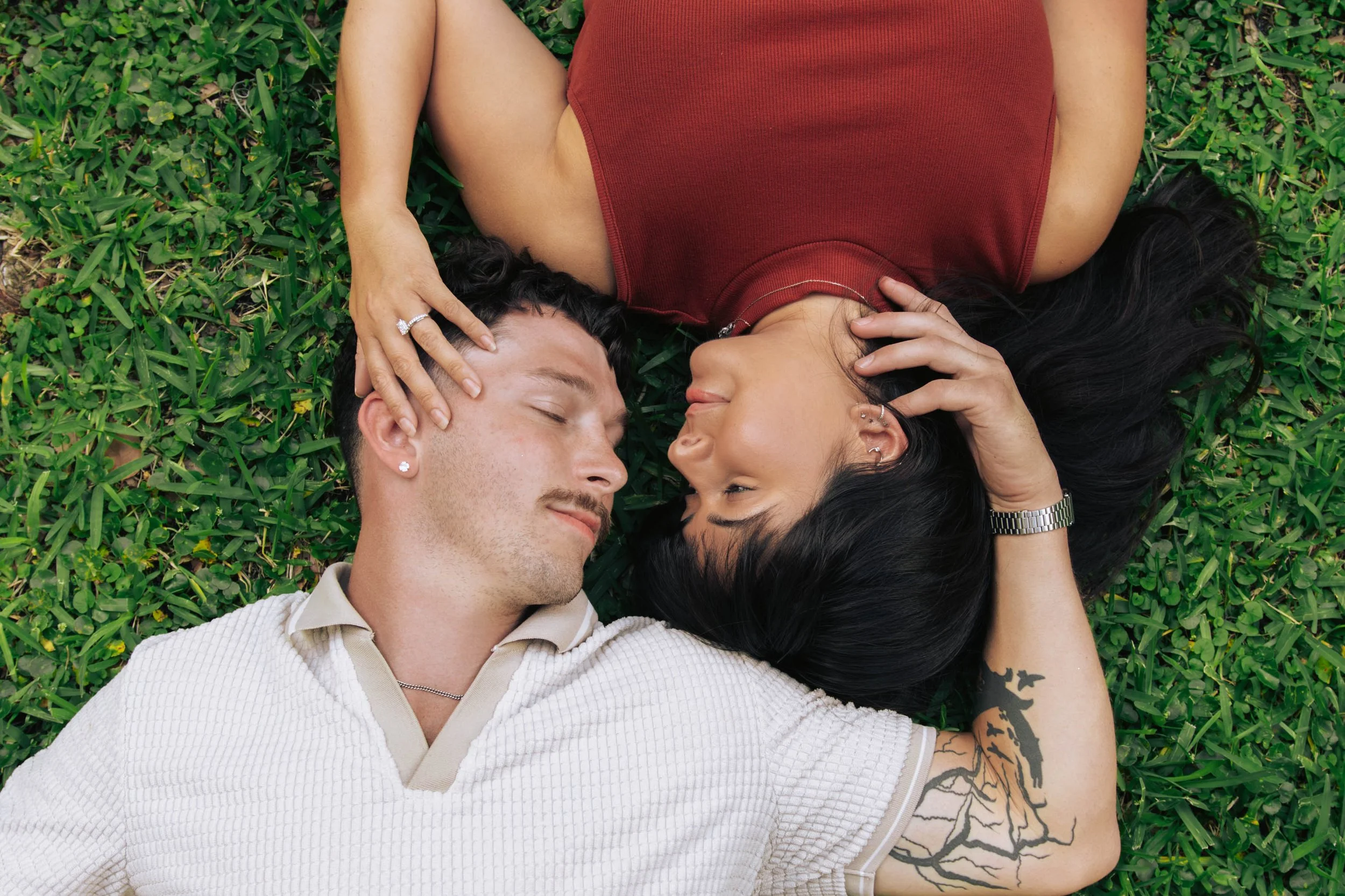 A couple lying on grass, the woman and man resting their heads near each other, smiling gently, with the woman touching the man's face.