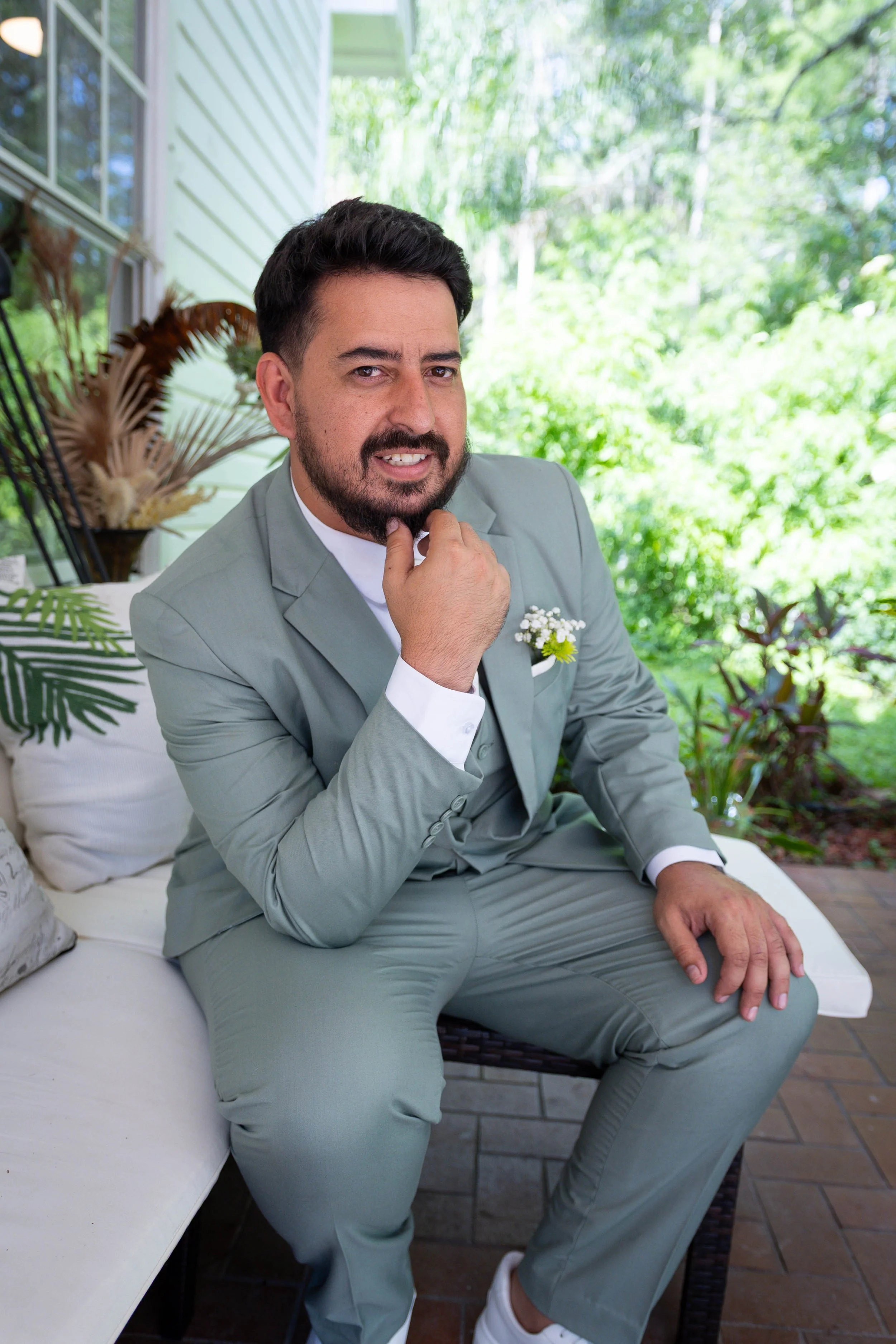 A man with dark hair, beard, and mustache in a light gray suit, sitting on a white outdoor sofa with greenery in the background, holding his chin and smiling.