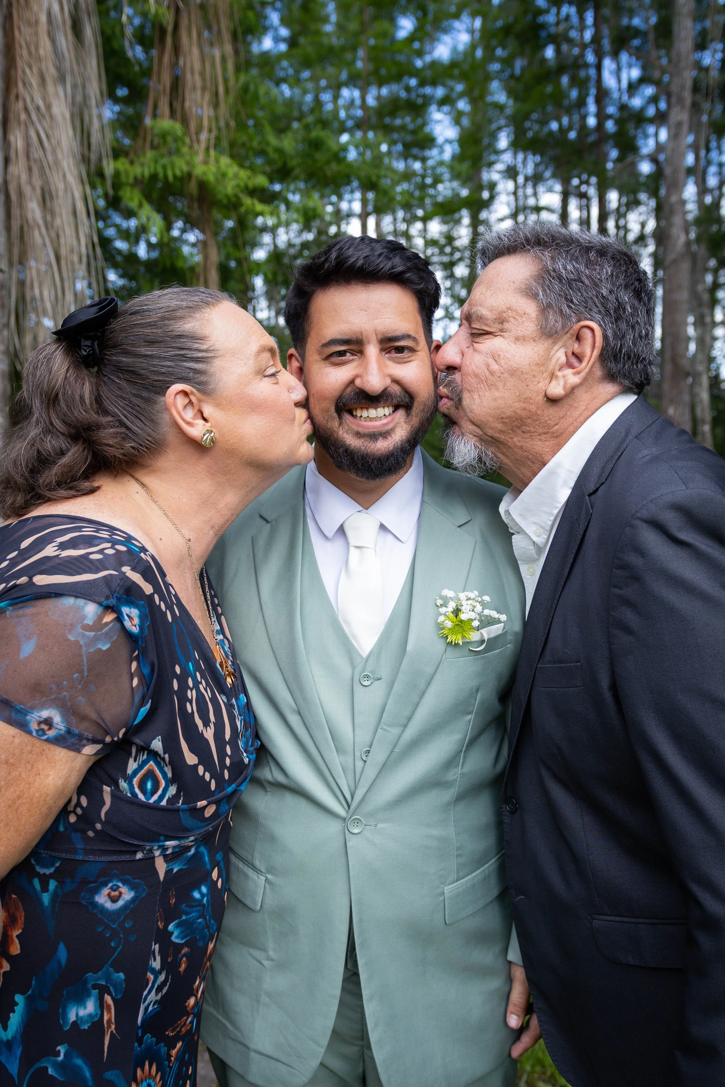 A groom is smiling as he is kissed on the cheeks by a woman and a man, possibly his parents, outside with green trees in the background.