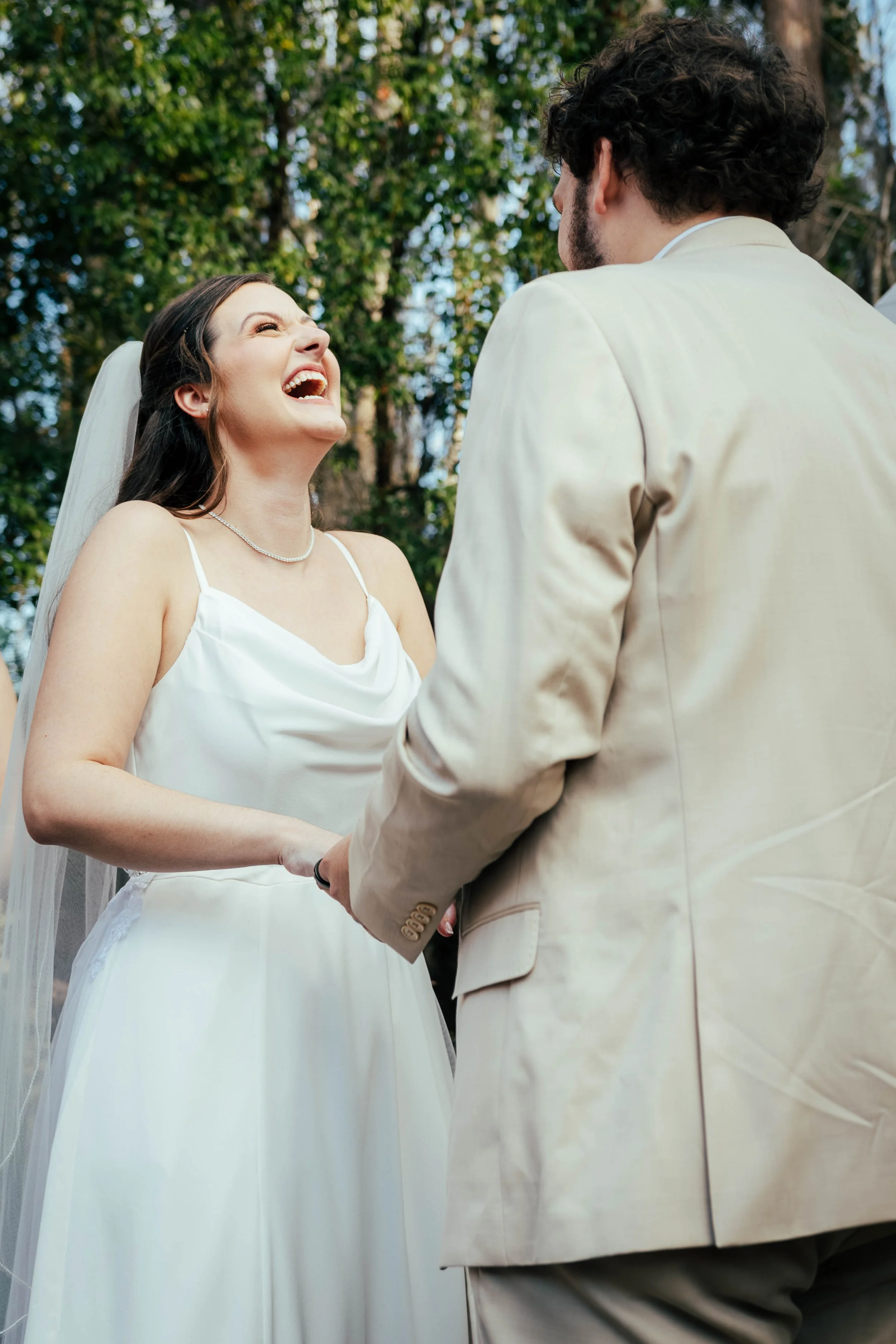 A woman and a man holding hands during a wedding ceremony outdoors, with green trees in the background.