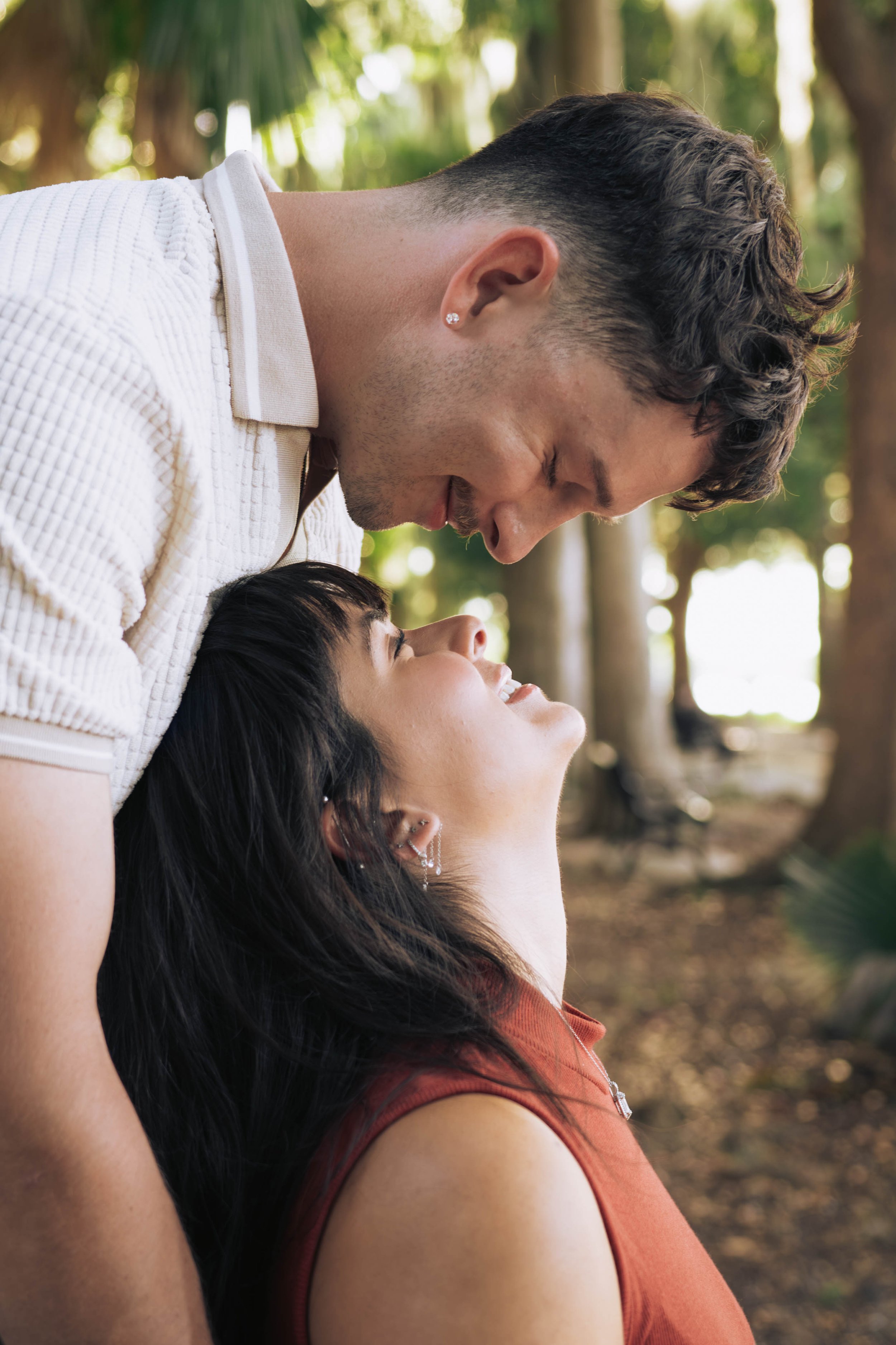A couple sharing an intimate moment outdoors, with the man leaning over the woman who is lying on her back in a natural setting, with trees and sunlight in the background.
