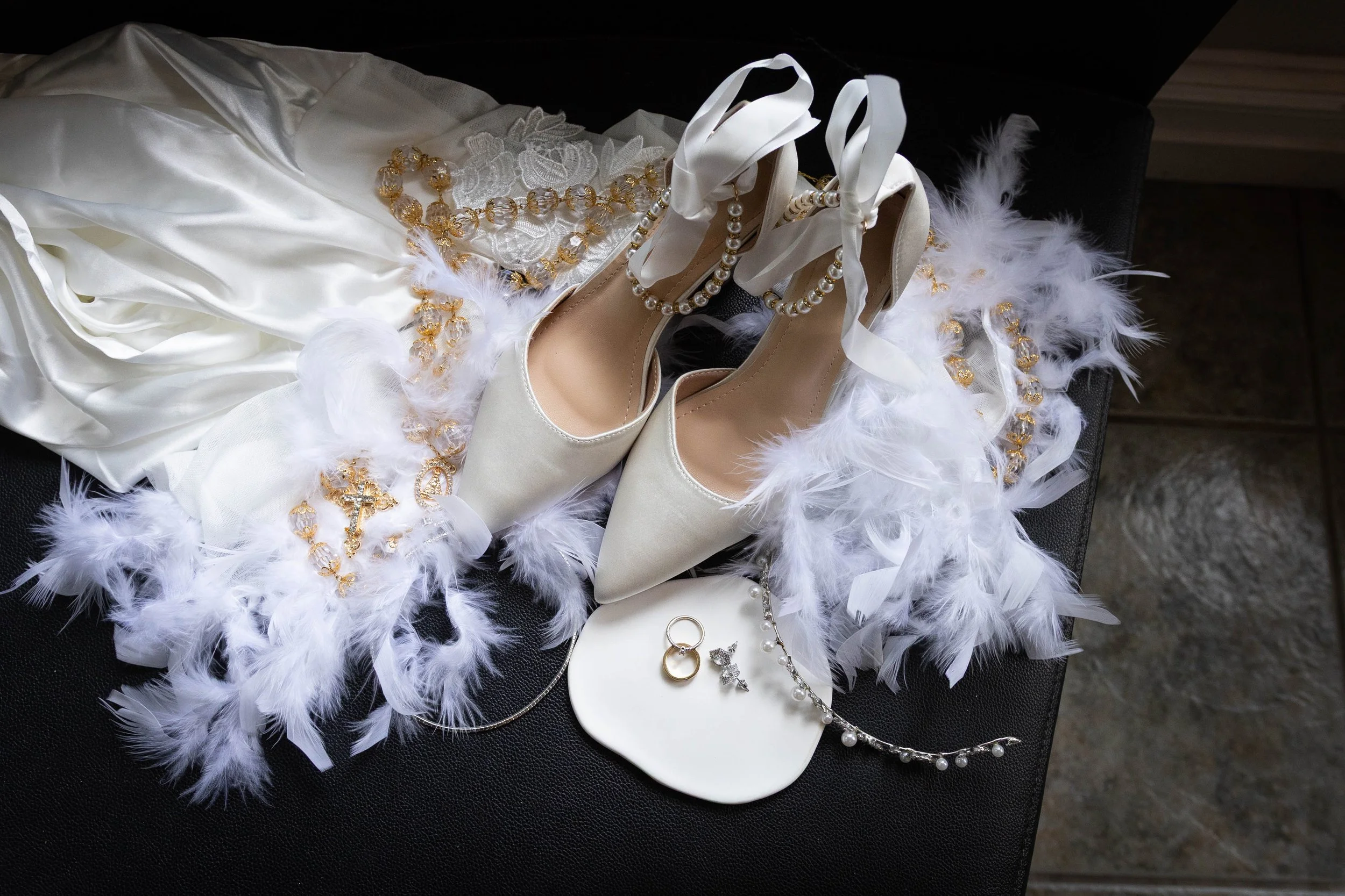 Wedding accessories including white satin shoes, pearl jewelry, lace gloves, and a satin dress with feather details.
