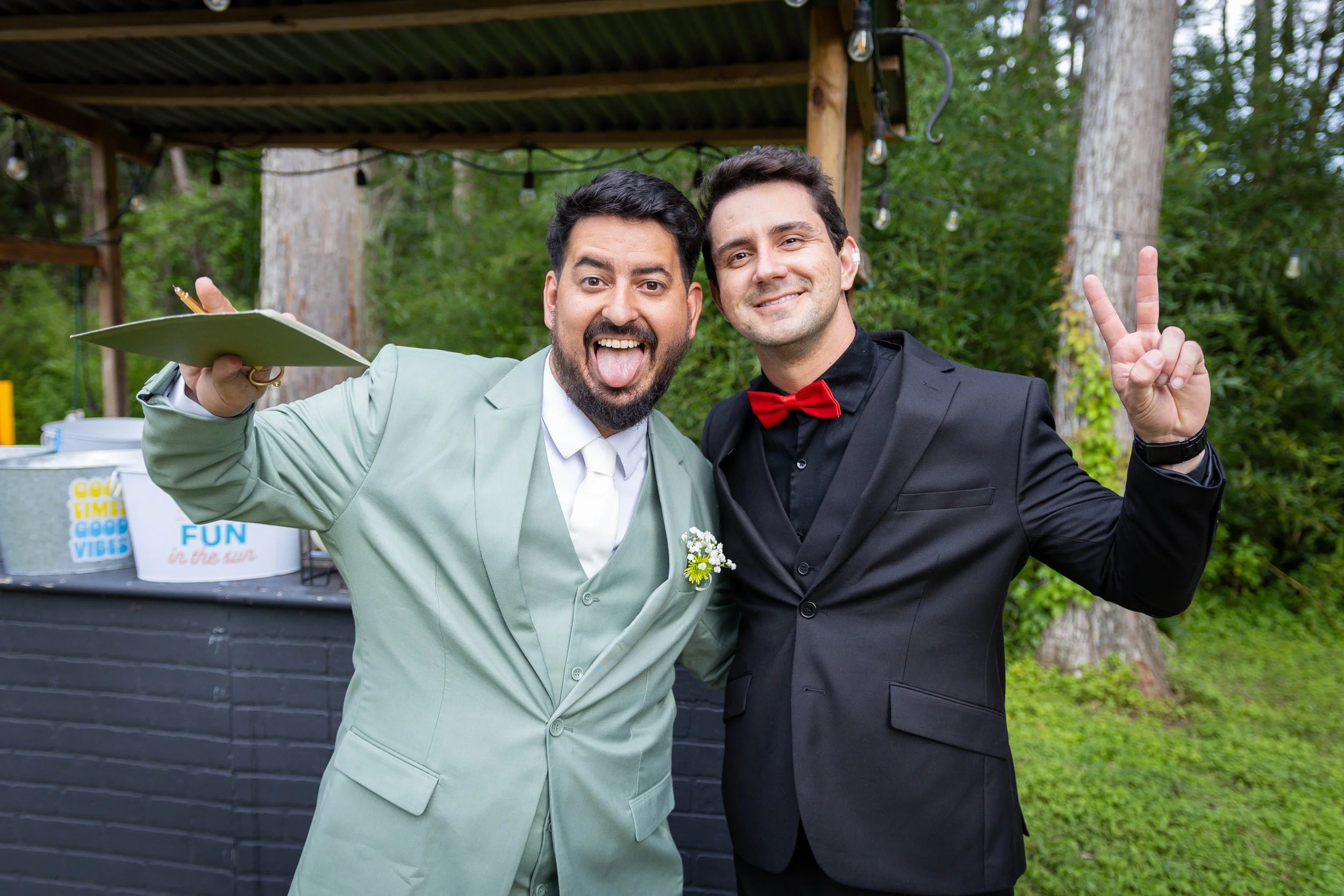 Two men dressed in suits smiling and posing for a photo at an outdoor event, one man making a peace sign and the other sticking out his tongue, with green trees and string lights in the background.