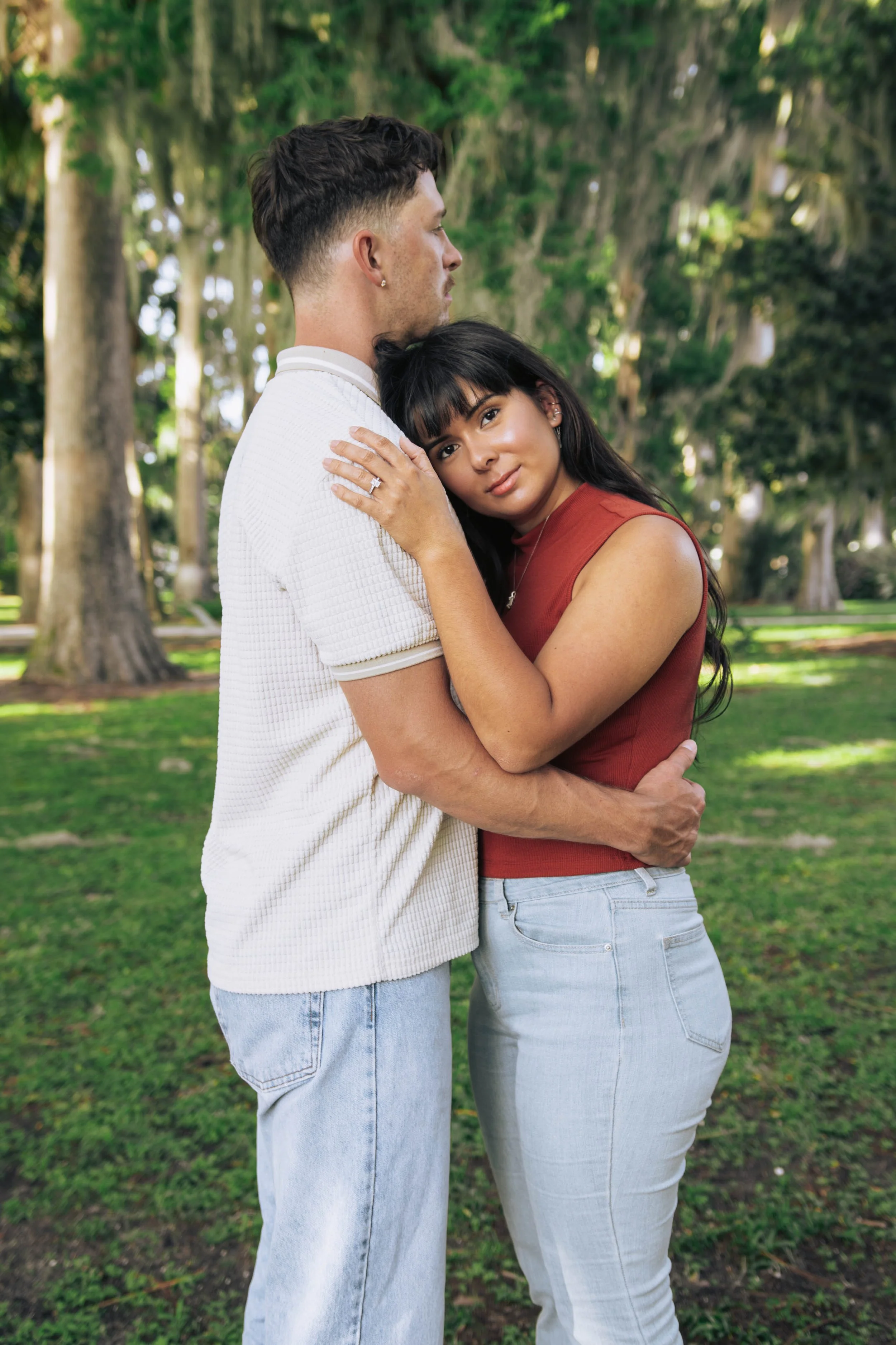 A man and a woman hugging outdoors in a park with trees in the background. The woman is resting her head on the man's shoulder, and the man is holding her with both arms.
