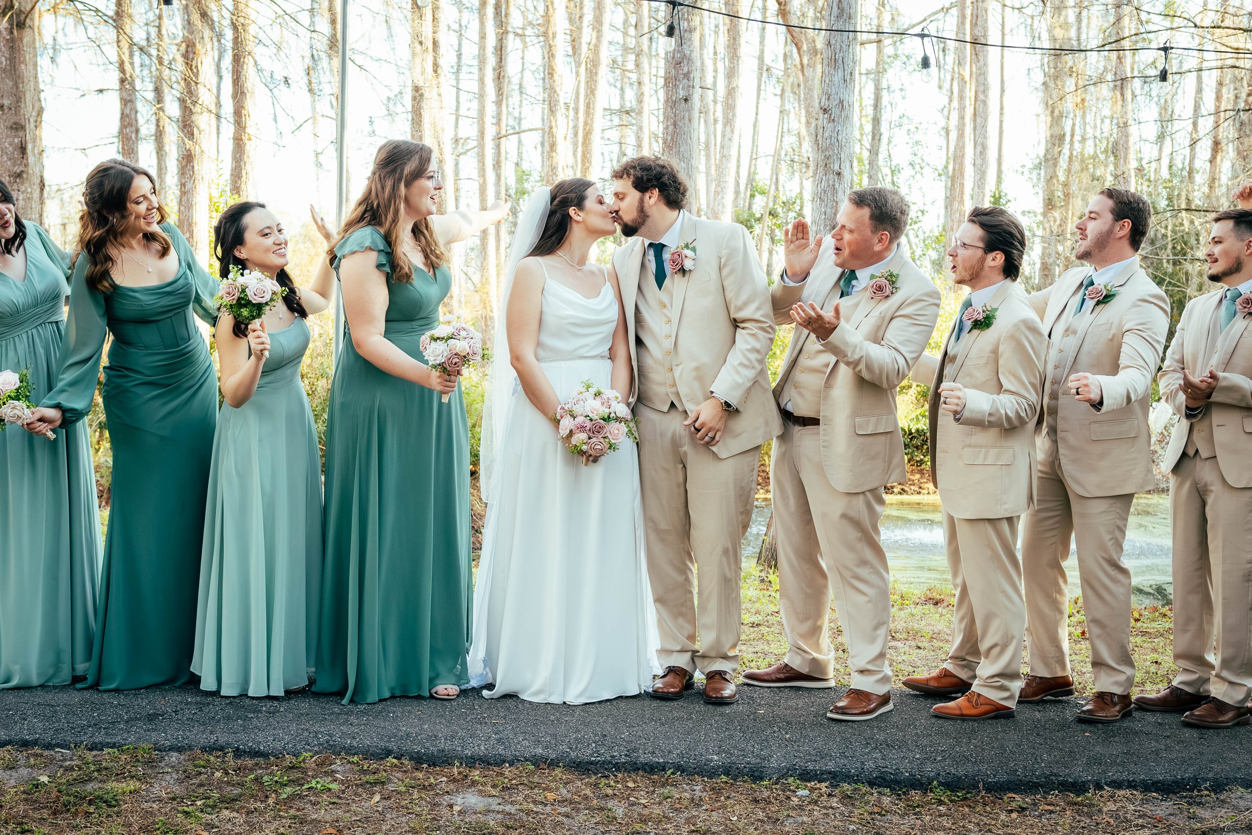 Bride and groom kiss while officiant and wedding party look on in an outdoor forest setting, with bridesmaids in teal dresses and groomsmen in beige suits standing nearby.