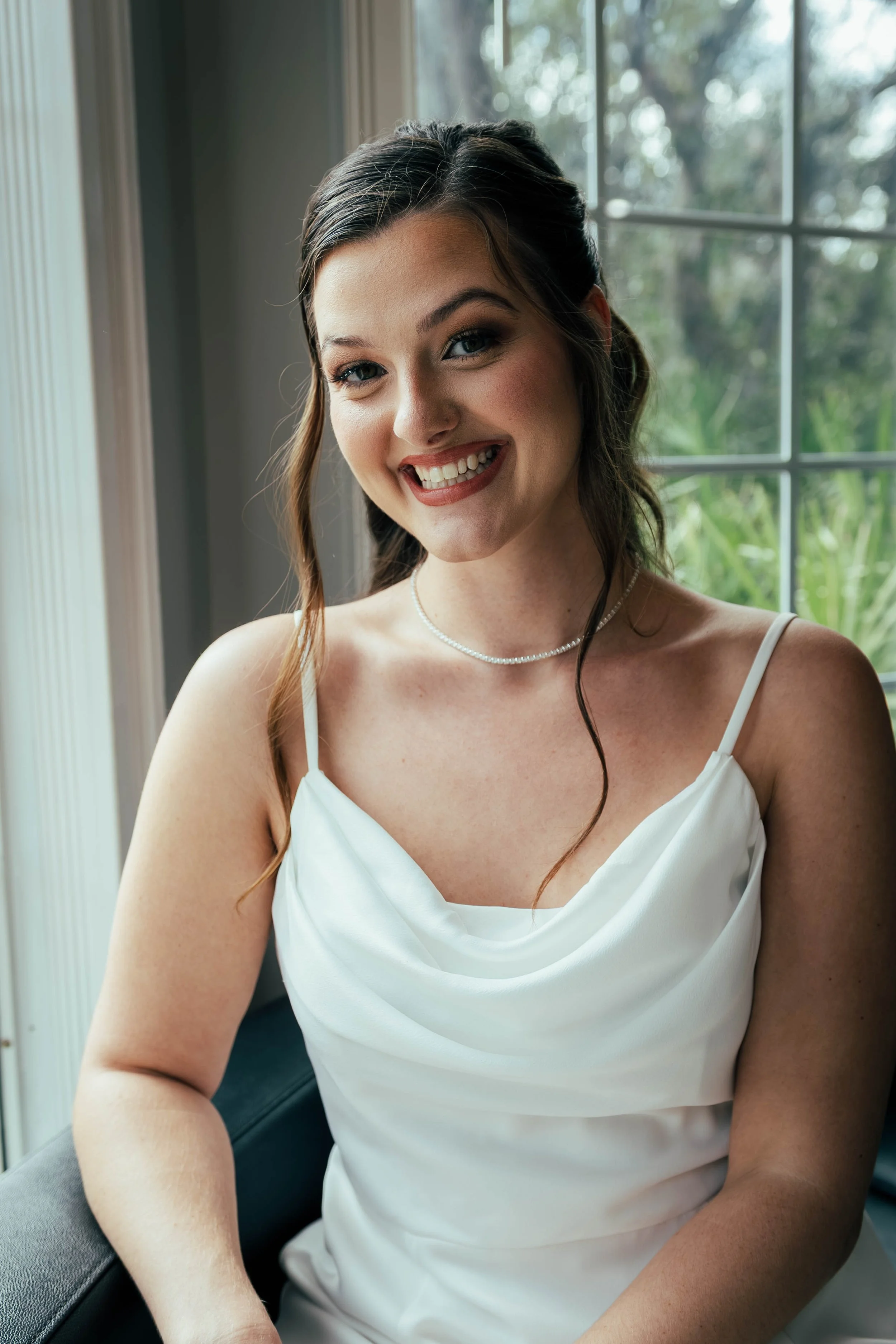 A woman smiling, sitting indoors near a window with a blurred outdoor view, wearing a white dress with thin straps, and a pearl necklace.