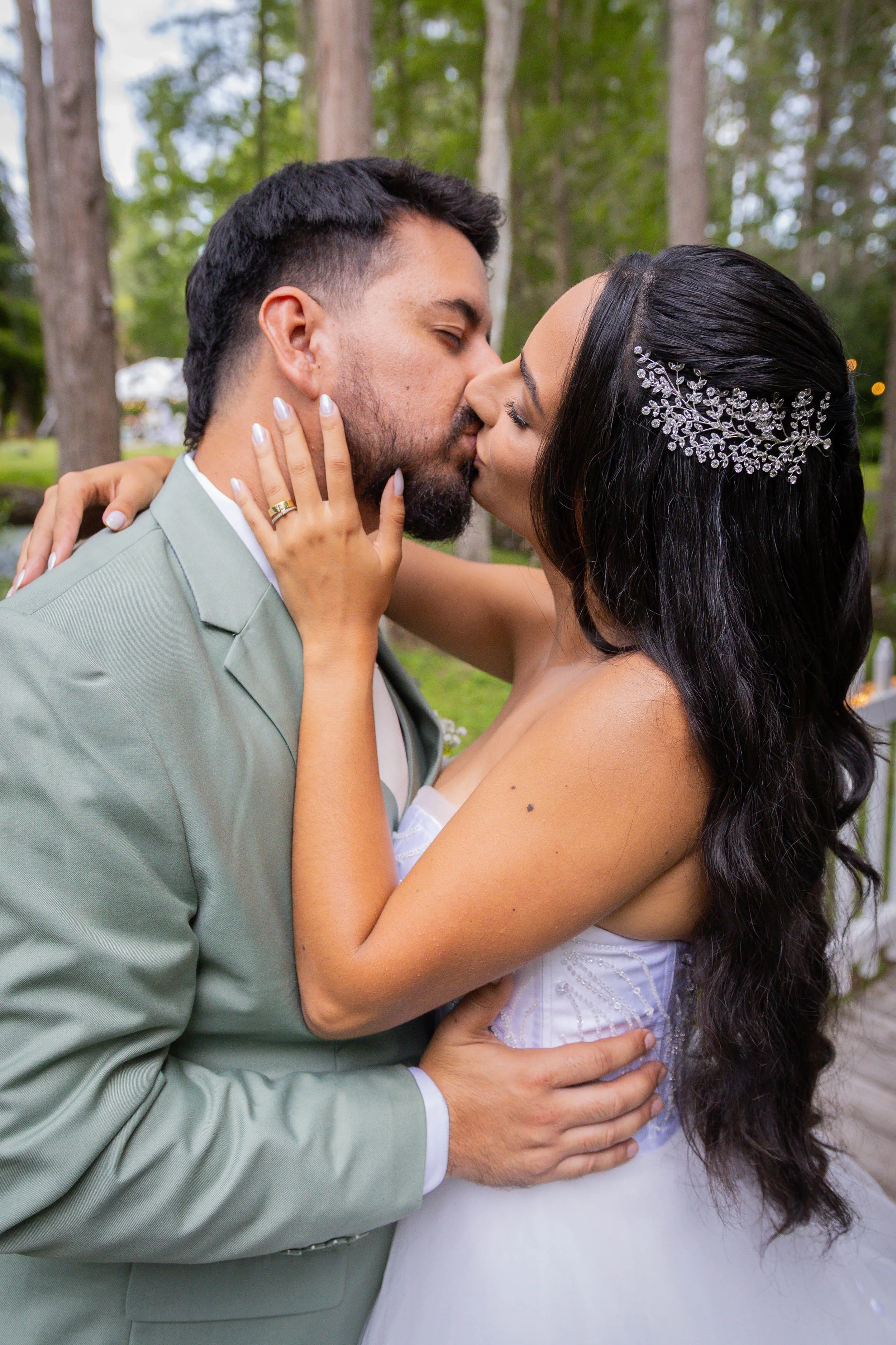 A couple sharing a kiss outdoors, with the woman wearing a white dress and a rhinestone hair accessory, and the man in a light green suit, surrounded by trees.