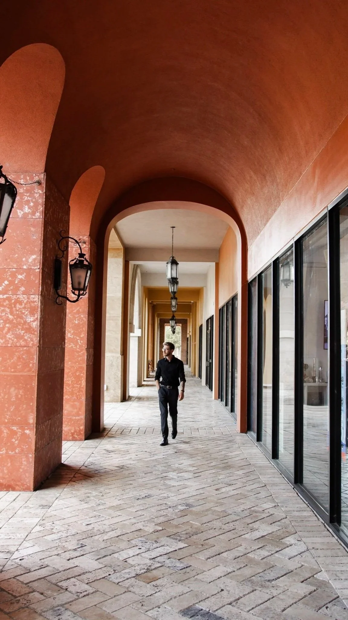A man in a black shirt and gray pants walking through a covered outdoor walkway with decorative lanterns, arched ceiling, and large glass windows reflecting the exterior.