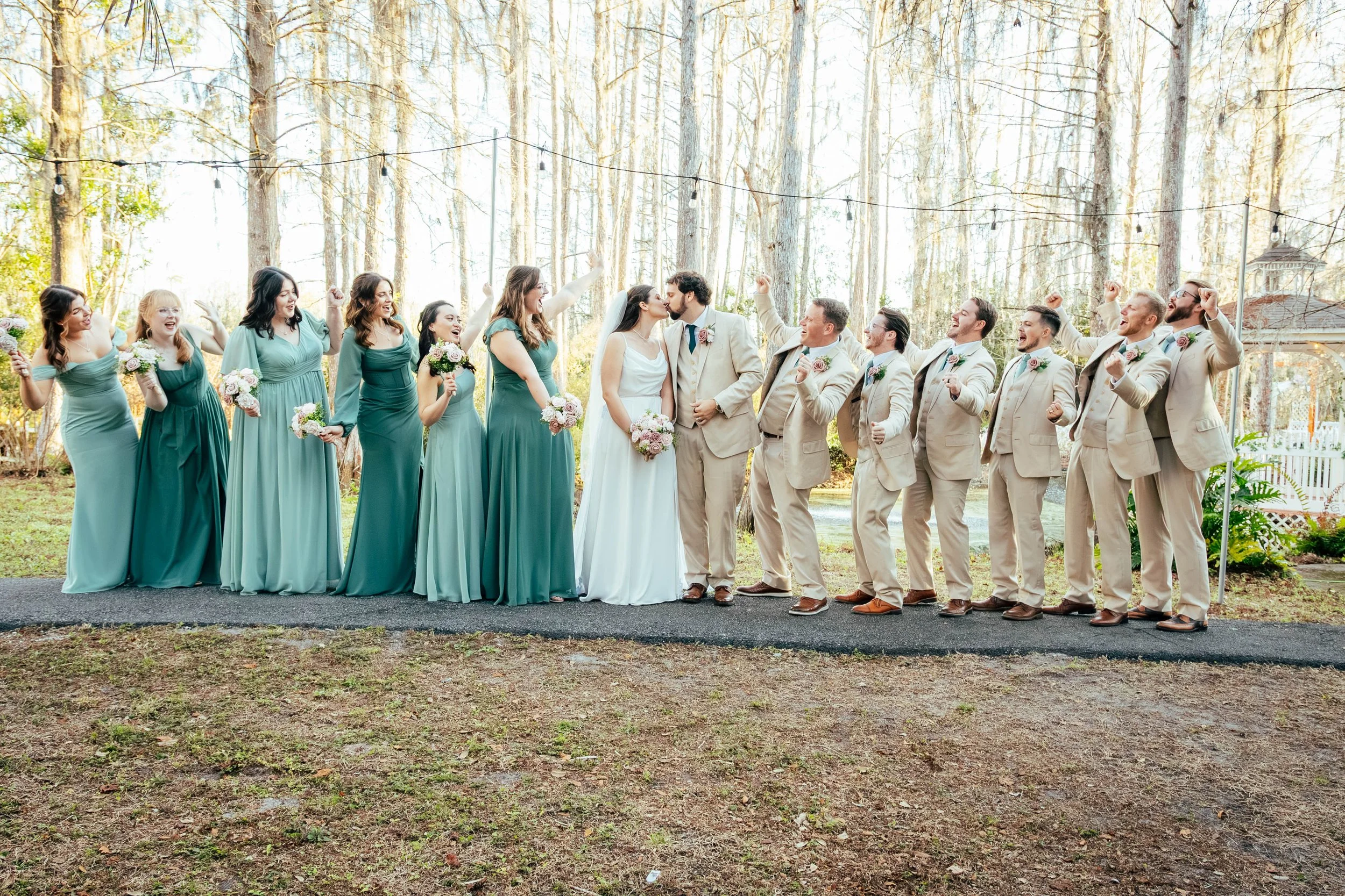 A wedding party standing outdoors in a wooded area with string lights overhead. The bride and groom are kissing at the center, surrounded by bridesmaids and groomsmen celebrating.