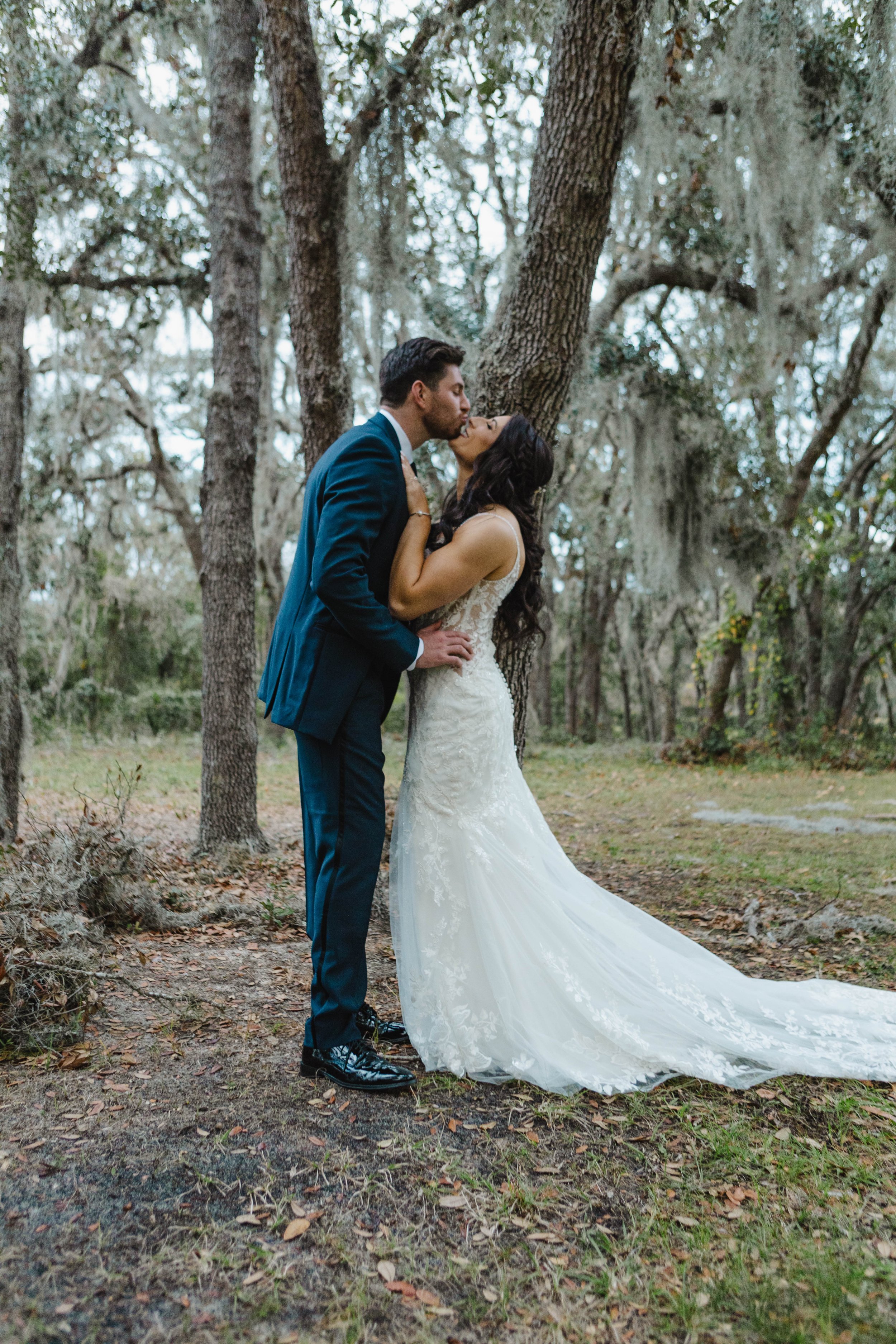 A bride and groom sharing a kiss in a wooded outdoor setting. The bride is wearing a white lace wedding gown with a train, and the groom is dressed in a dark blue suit with black shoes.