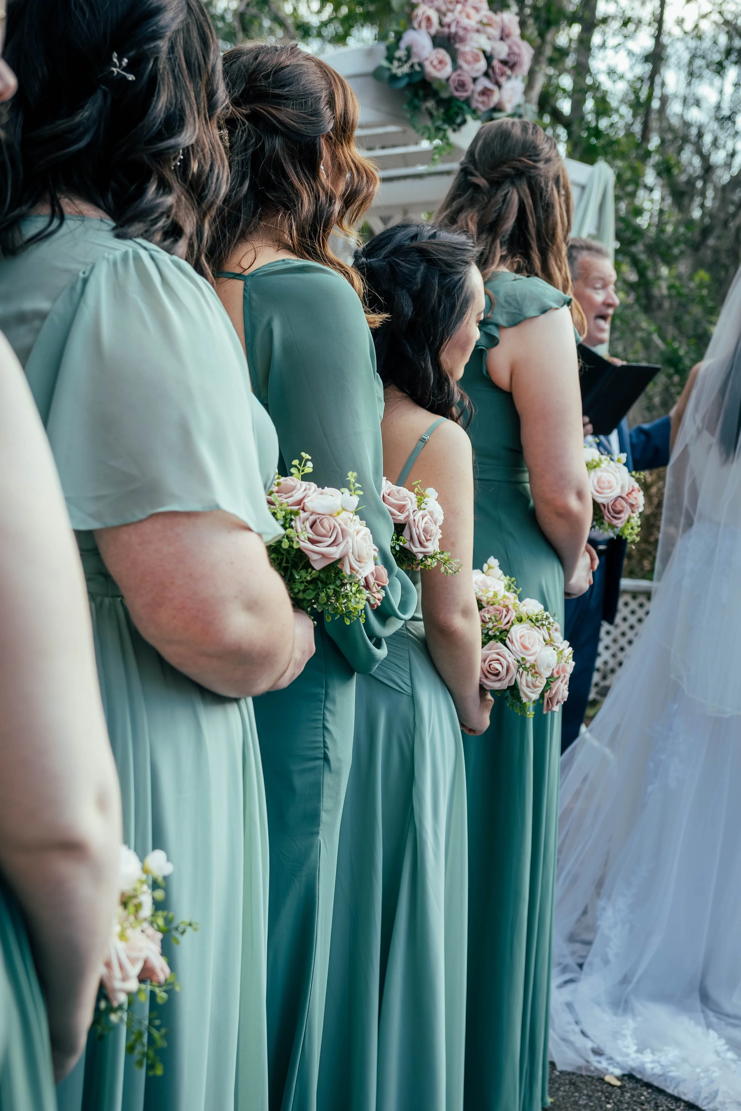 A lineup of women wearing matching light blue dresses, holding bouquets, at a wedding ceremony outdoors. The women are standing side by side with greenery in the background, and a man speaking to the bride can be seen on the right side.