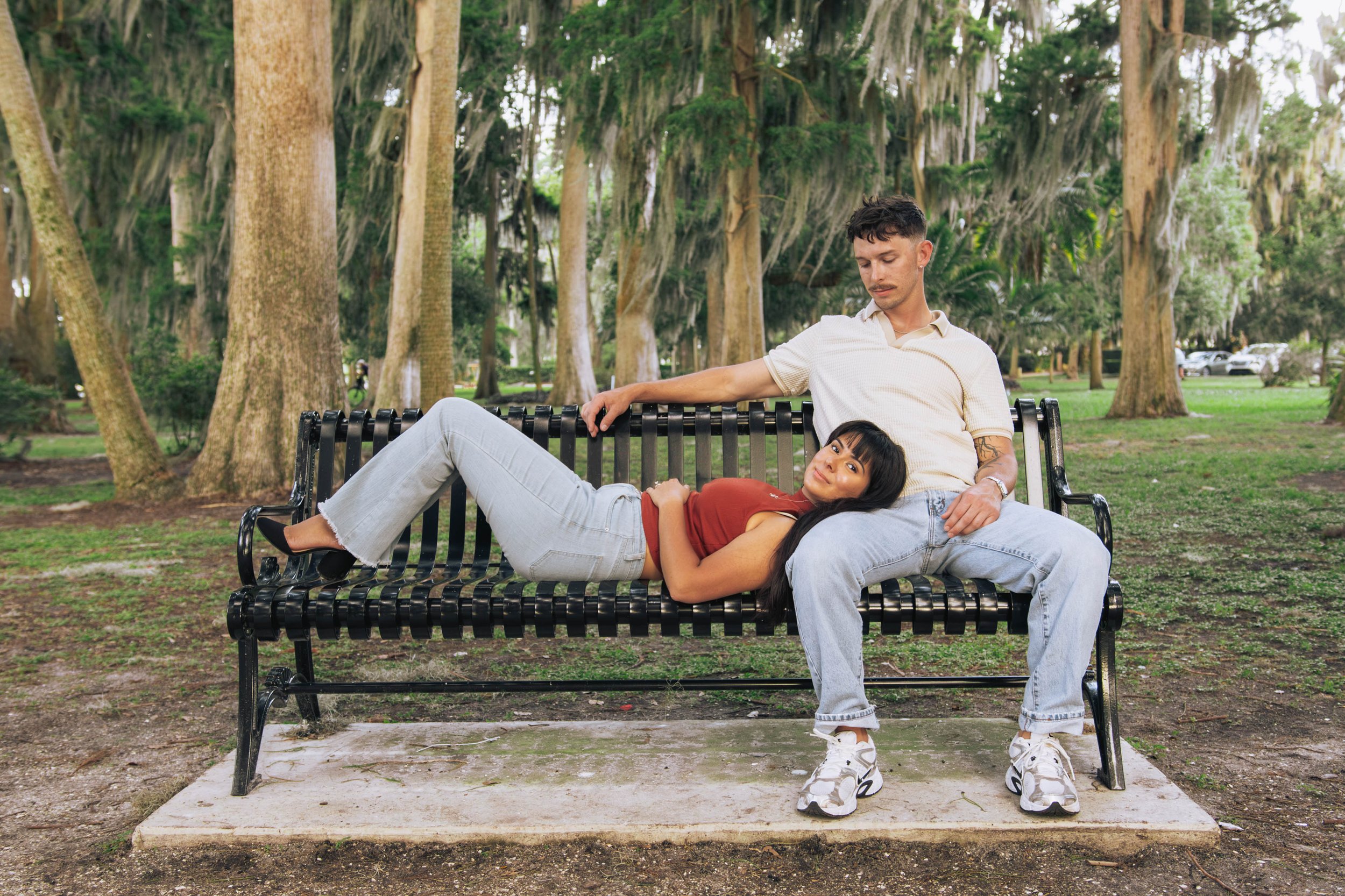 A young man and woman sitting on a park bench surrounded by tall trees with Spanish moss hanging from the branches. The woman is lying down with her head on the man's lap, and they are looking into each other's eyes.