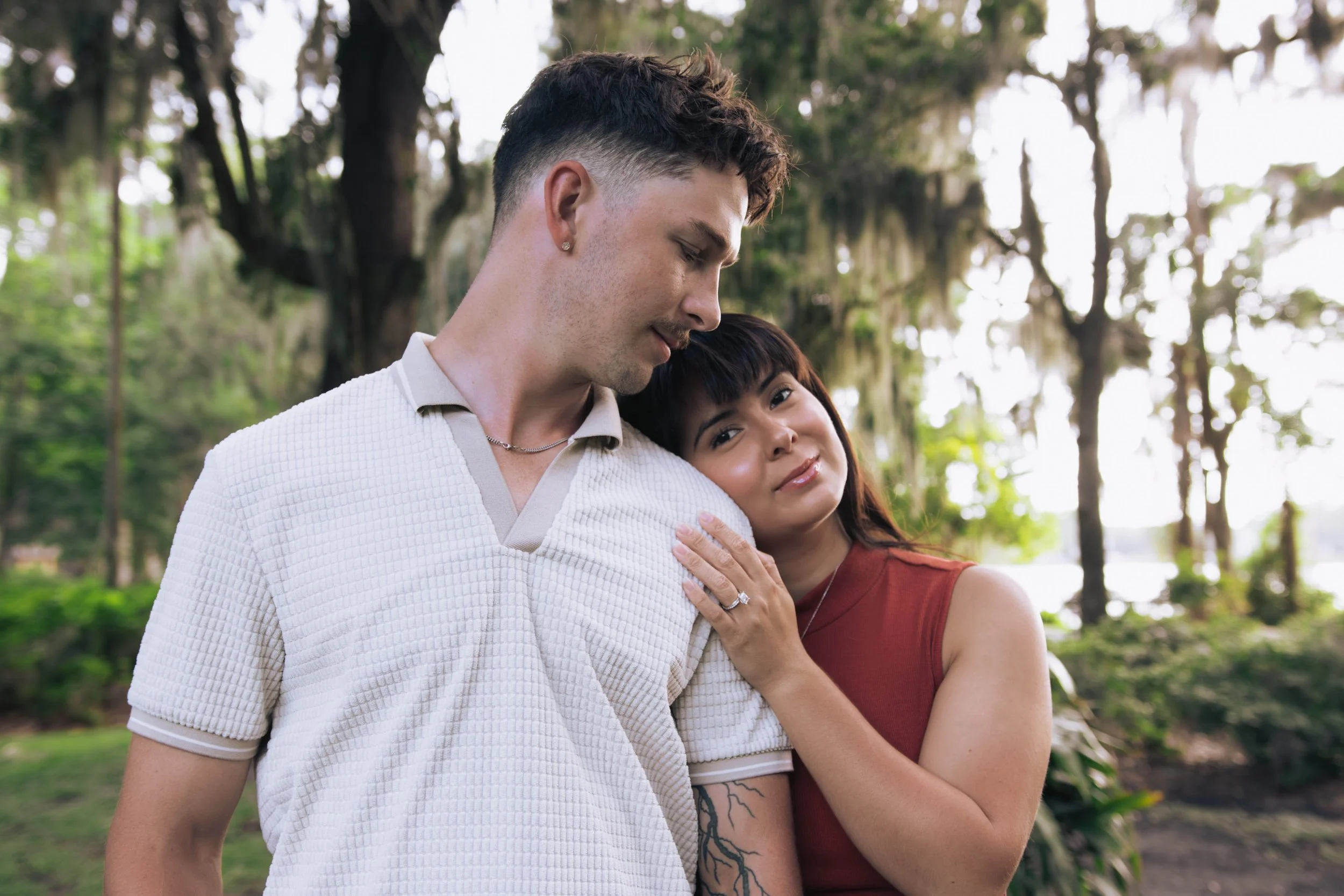 A young couple with light skin enjoying a moment together outdoors in a wooded area, with the woman resting her head on the man's shoulder.