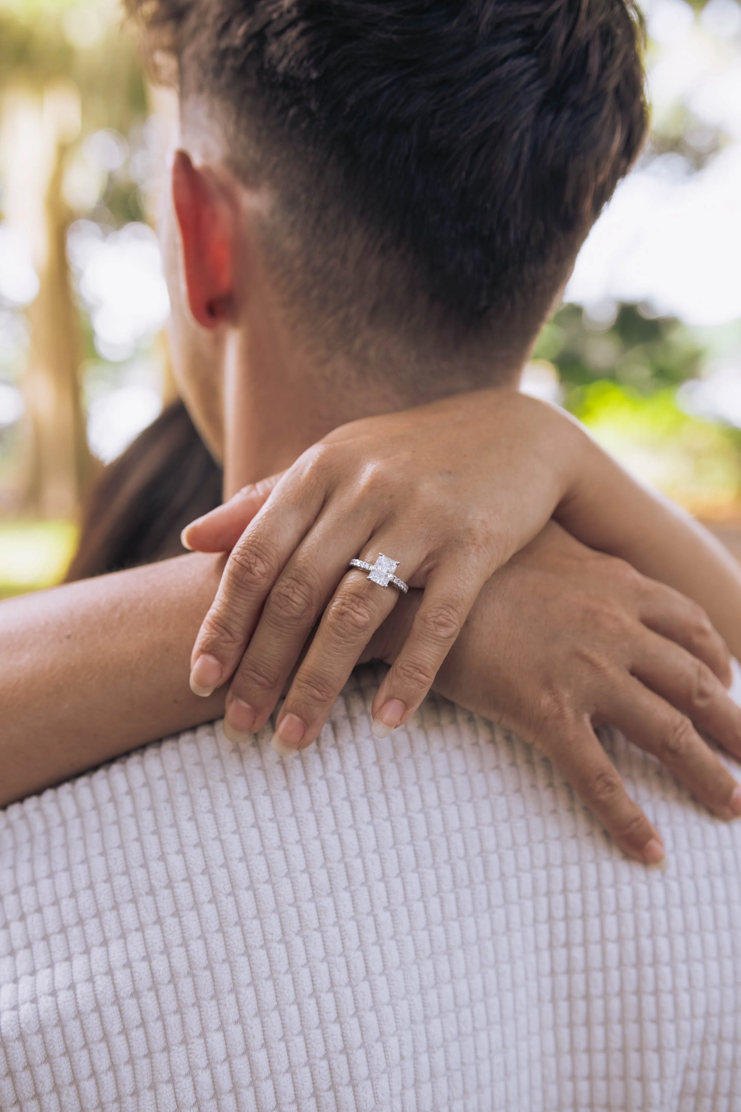Close-up of two people embracing, focusing on one person's hand with an engagement ring on their finger, against a blurred outdoor background.