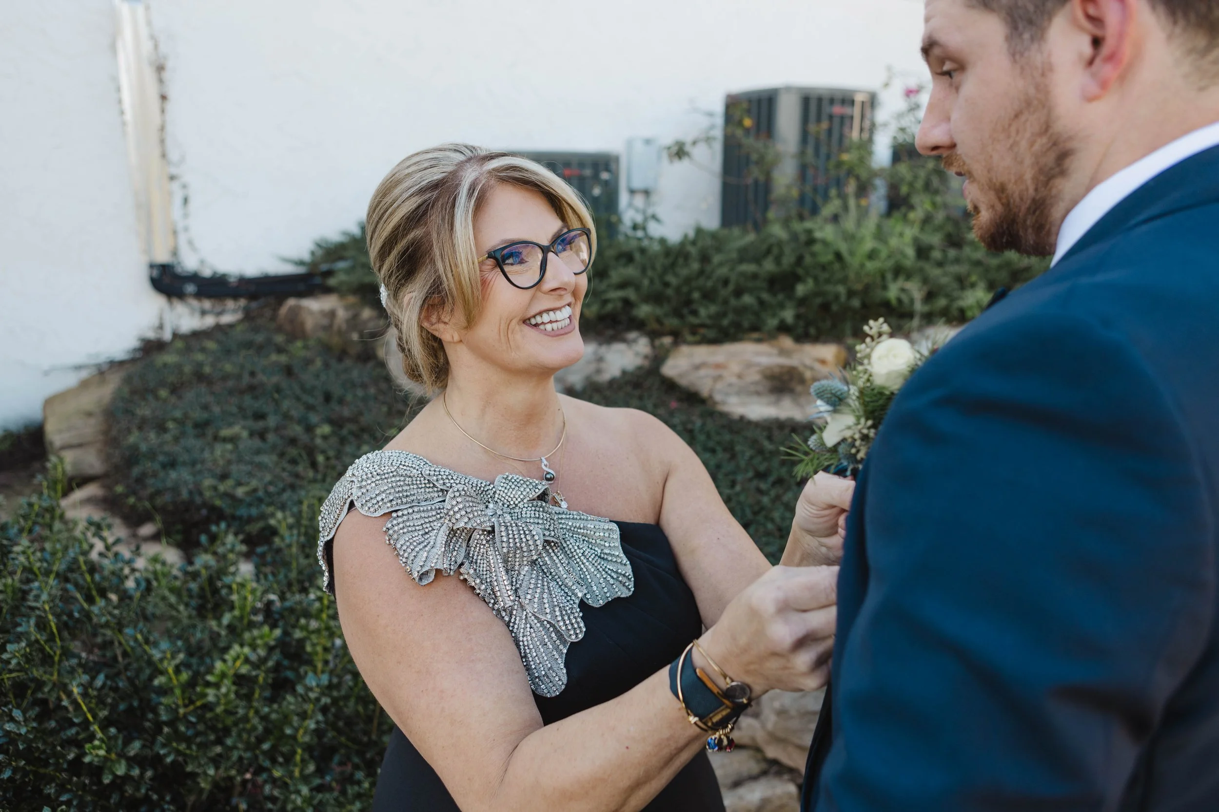 A woman with glasses smiling and holding hands with a man in a blue suit, outdoor setting with greenery and white wall.