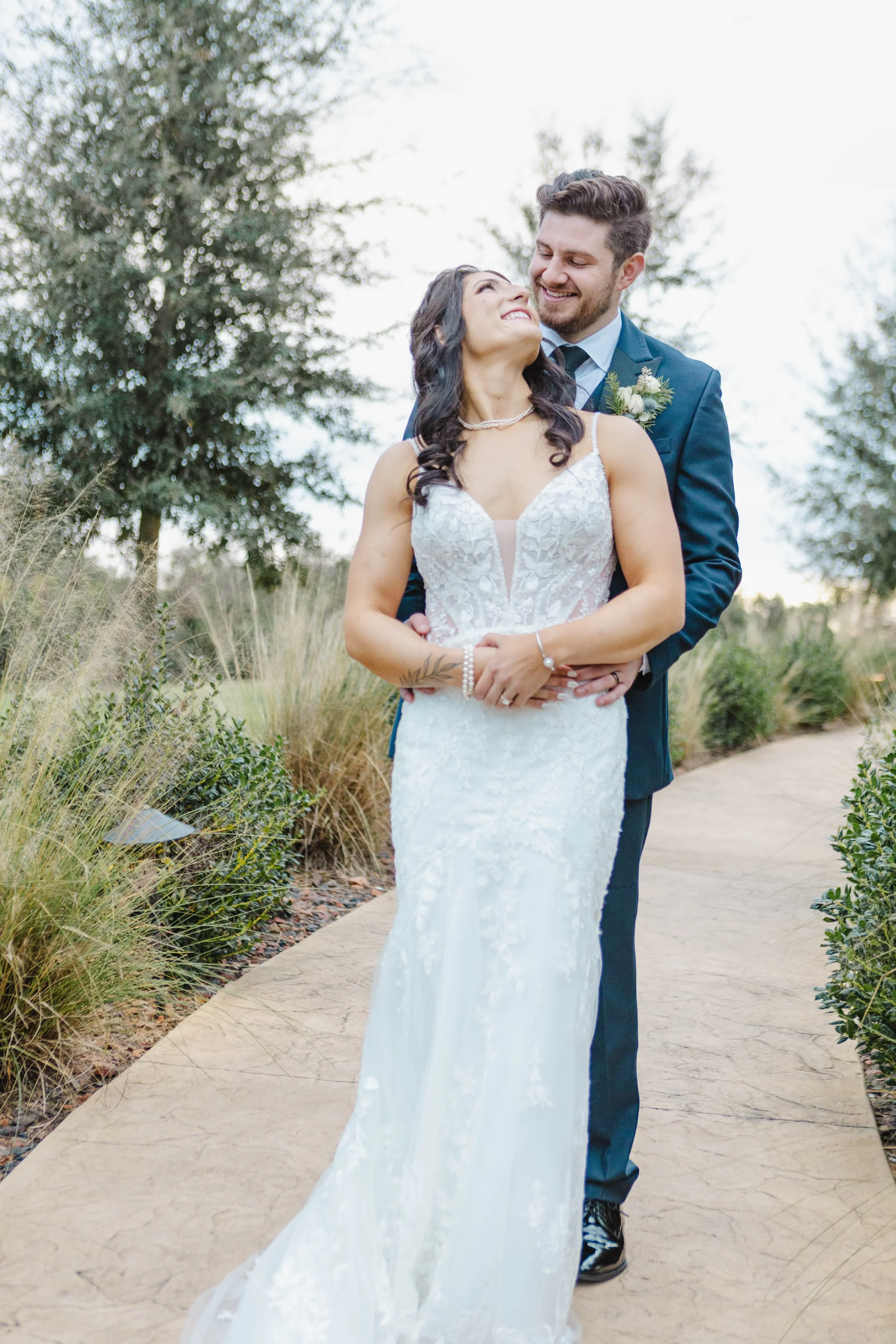 A smiling couple in wedding attire standing outdoors on a paved path surrounded by greenery and trees, with the woman in a white wedding gown and the man in a blue suit.