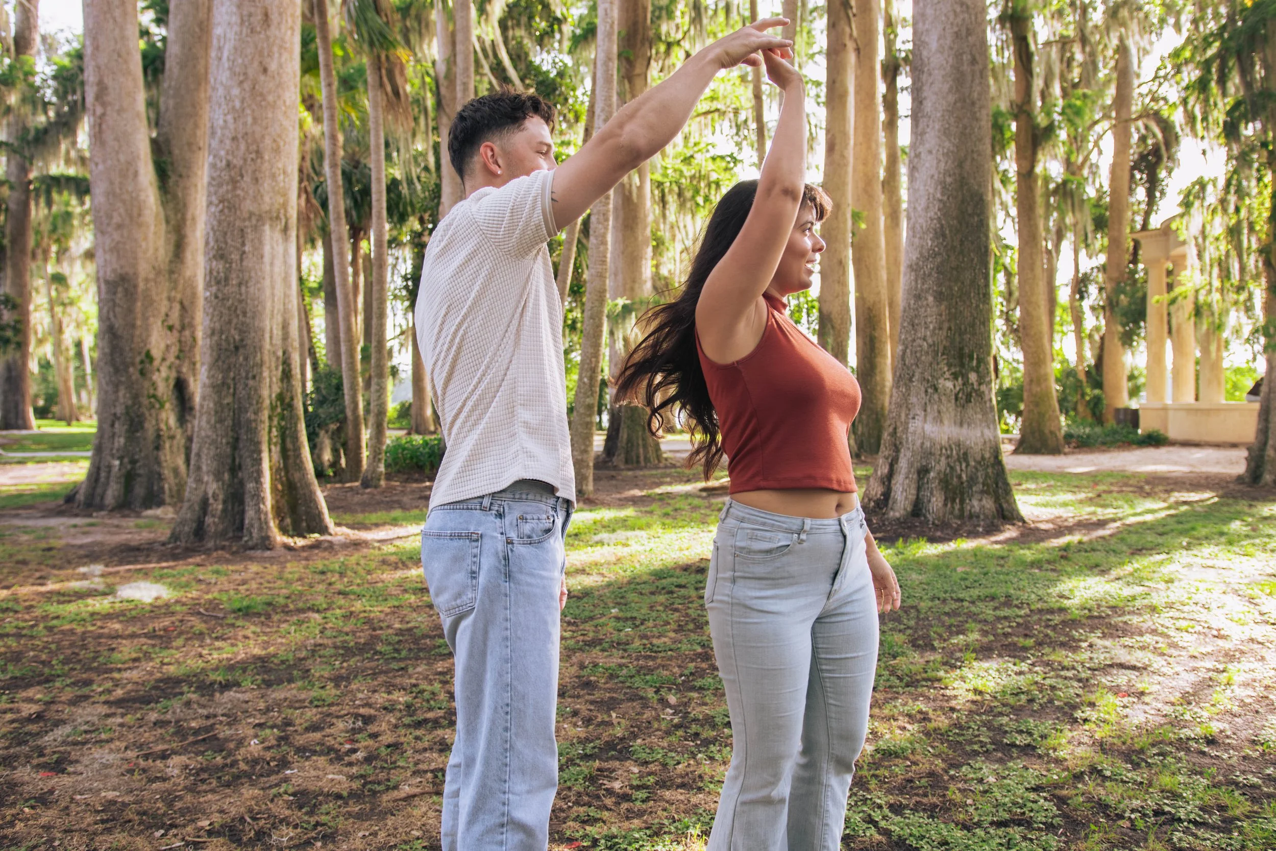 A man and woman stand outdoors in a park with tall trees, with the man raising his arms above the woman's head, both smiling and enjoying the moment.
