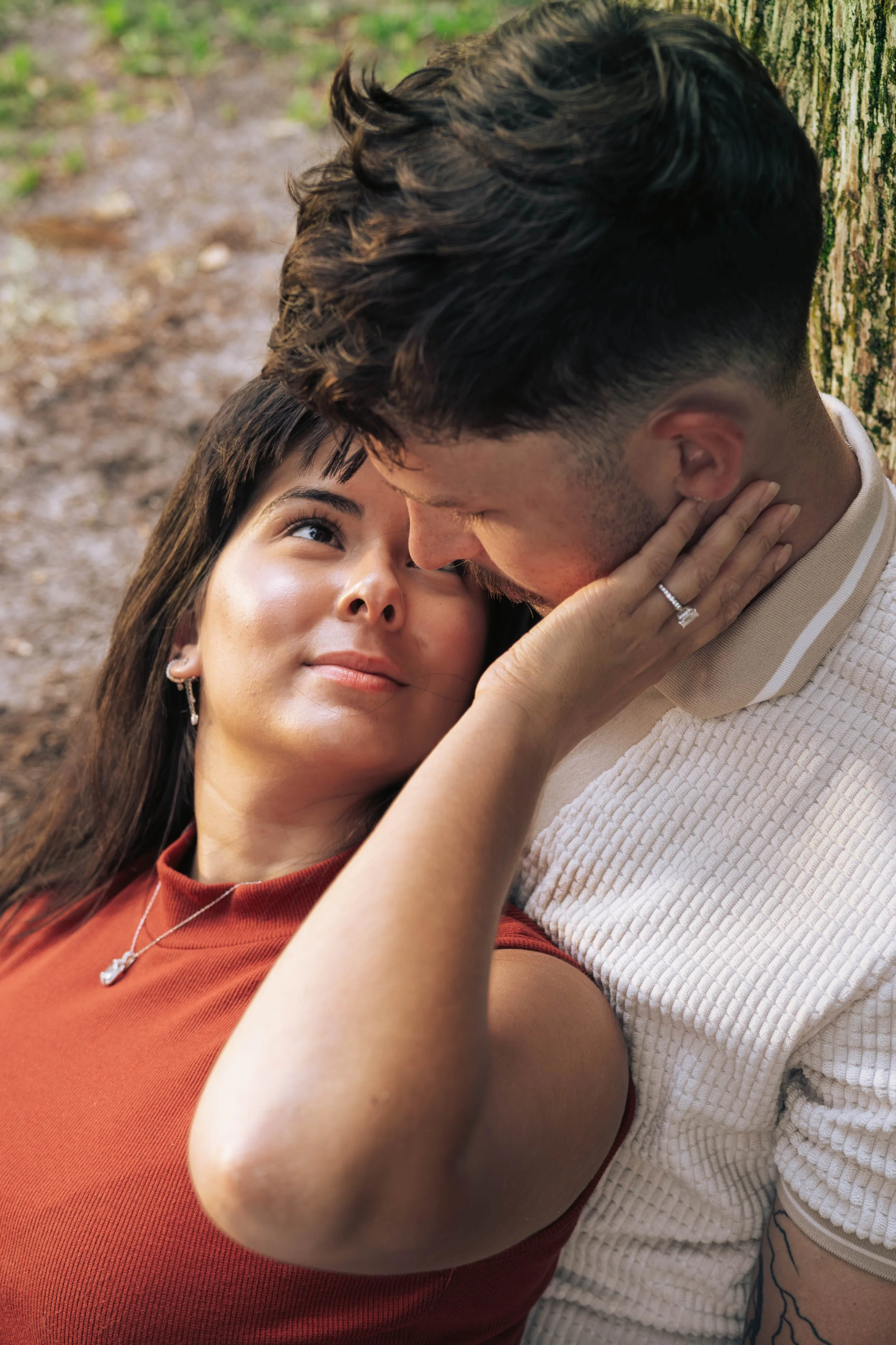 A couple sitting outdoors on the ground, gazing into each other's eyes, with the woman's hand on the man's face, showing her engagement ring.