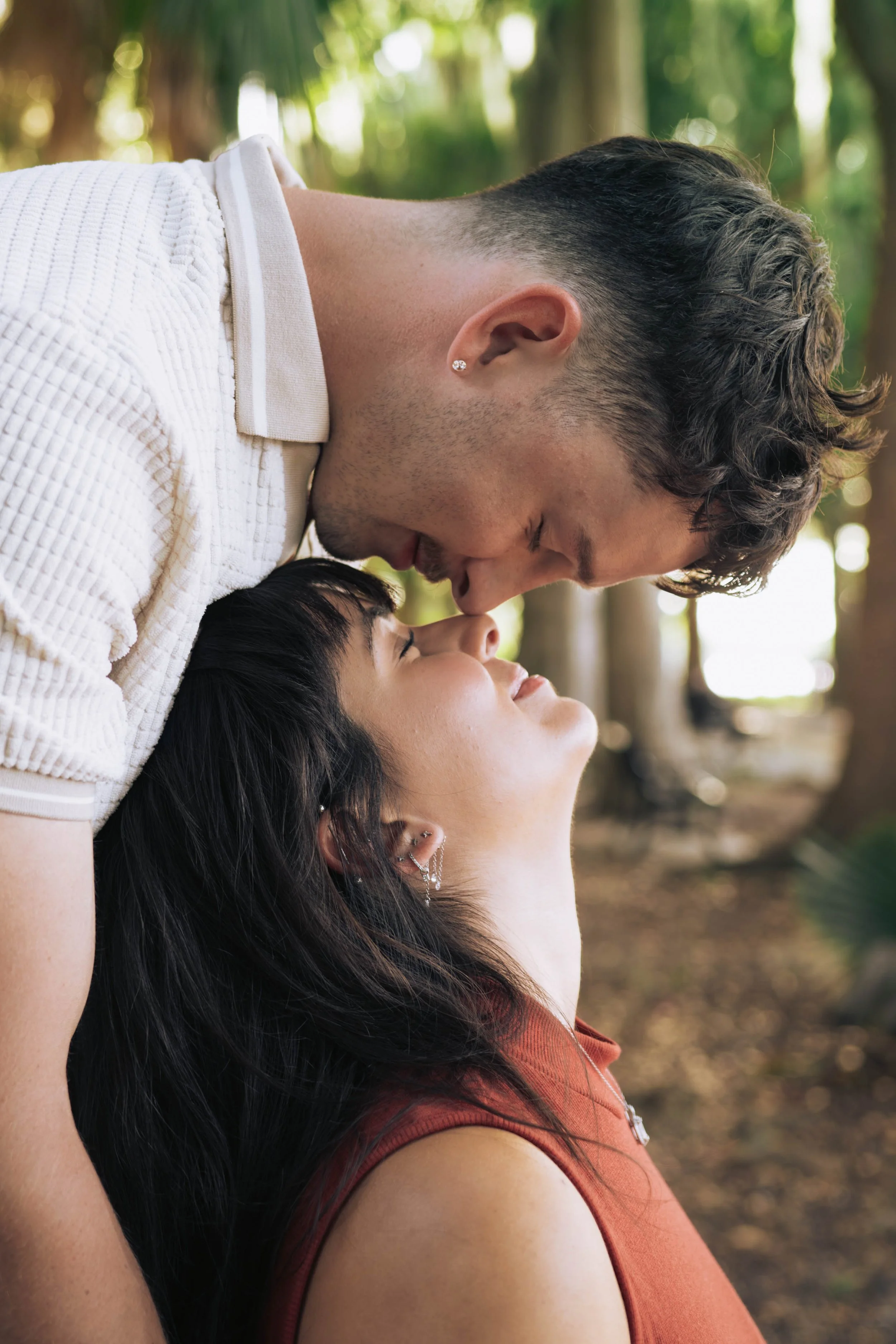 Two people sharing an intimate moment outdoors, the man leaning over the woman with their foreheads touching, surrounded by trees.