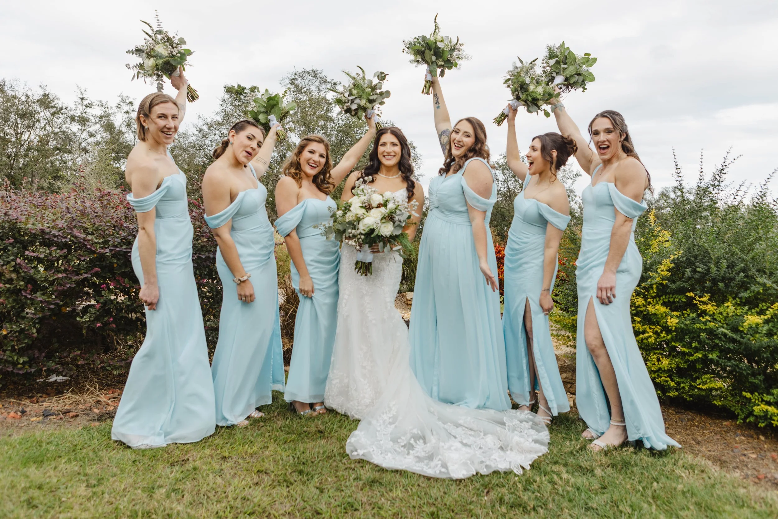 A group of seven women dressed in matching light blue dresses celebrating outdoors, with the bride in a white wedding gown holding a bouquet of flowers and others raising bouquets in the air, smiling and cheerful.