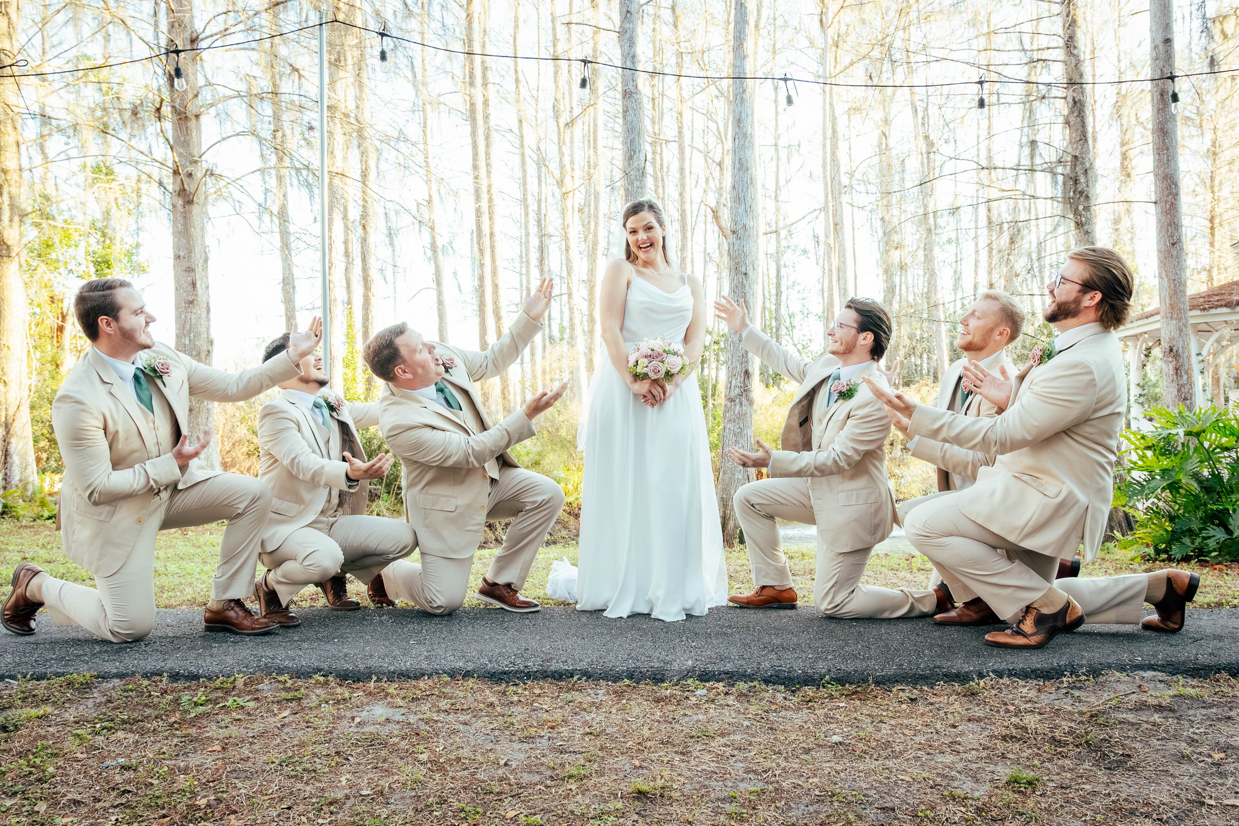 A bride in a white dress holding a bouquet surrounded by six groomsmen in beige suits kneeling on one knee with one hand raised in front of a wooded outdoor setting.