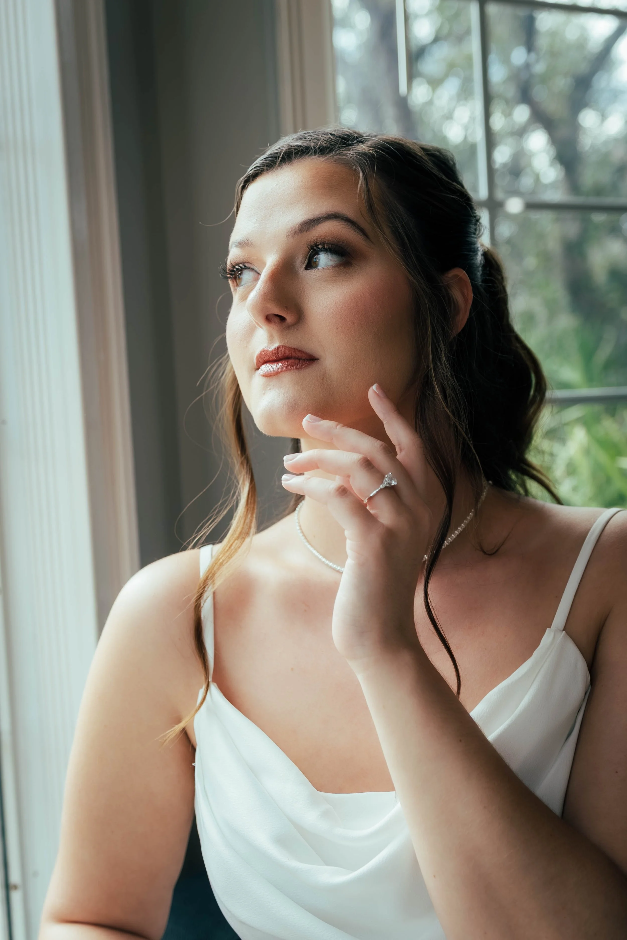 A woman with long brown hair styled in soft waves, wearing a white dress with thin straps. She is looking out a window with greenery outside, touching her chin with her right hand, showcasing a ring on her finger. The natural light from the window hi