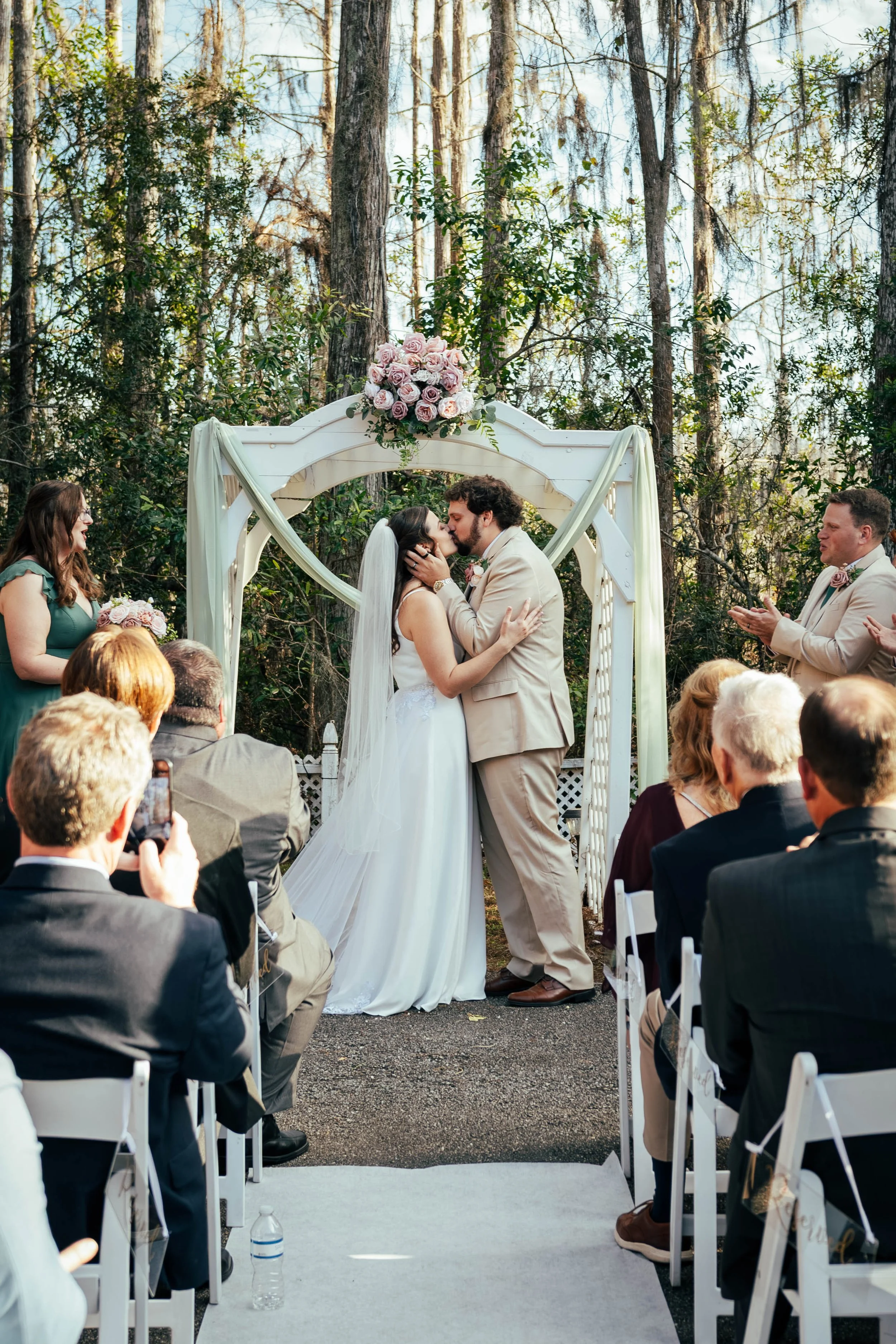 A couple sharing a kiss during their outdoor wedding ceremony with guests watching, officiant on the side, and a decorative floral arch behind them in a wooded area.