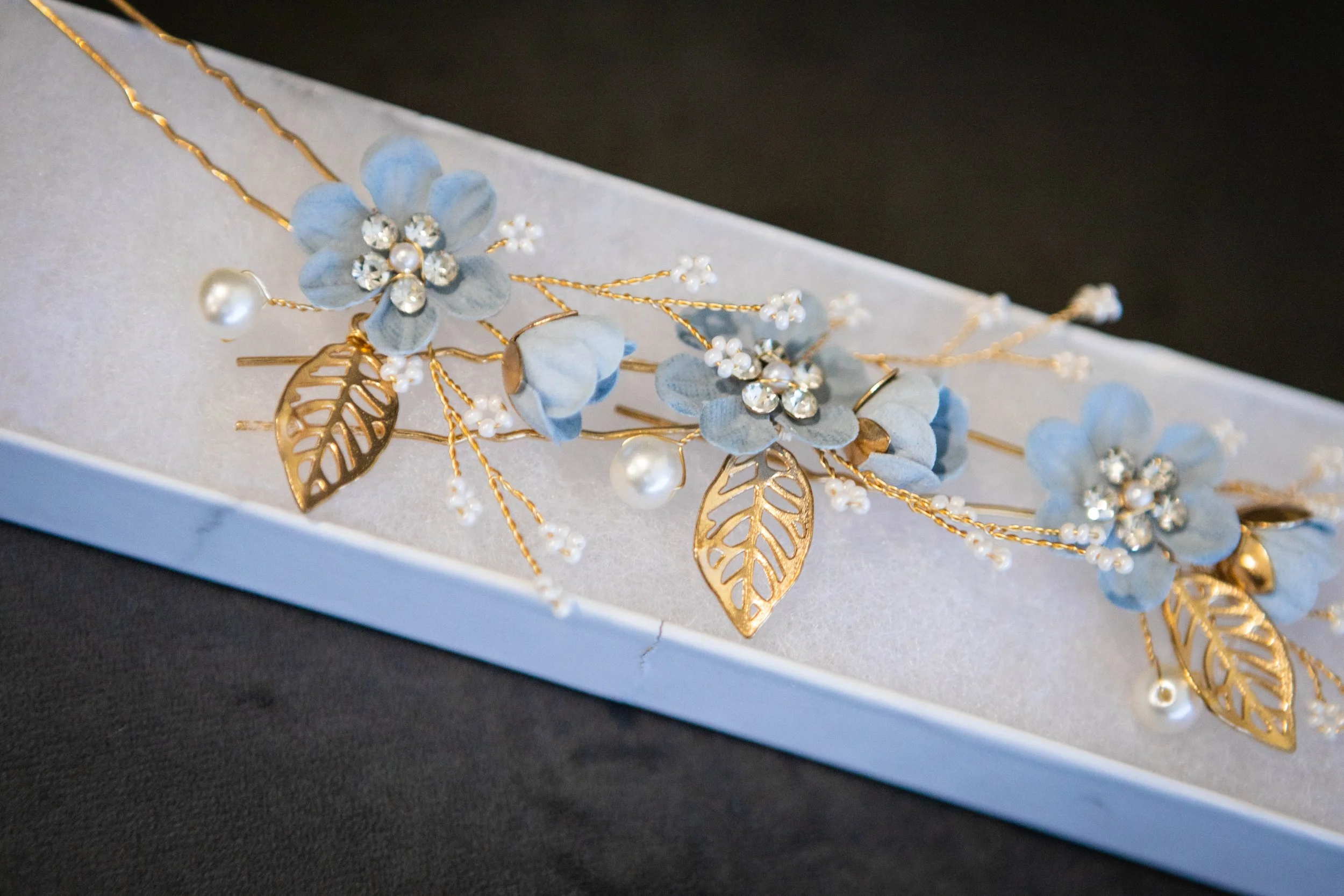 Gold floral headpiece with blue flowers, pearl accents, and leaf details, displayed on a white box.