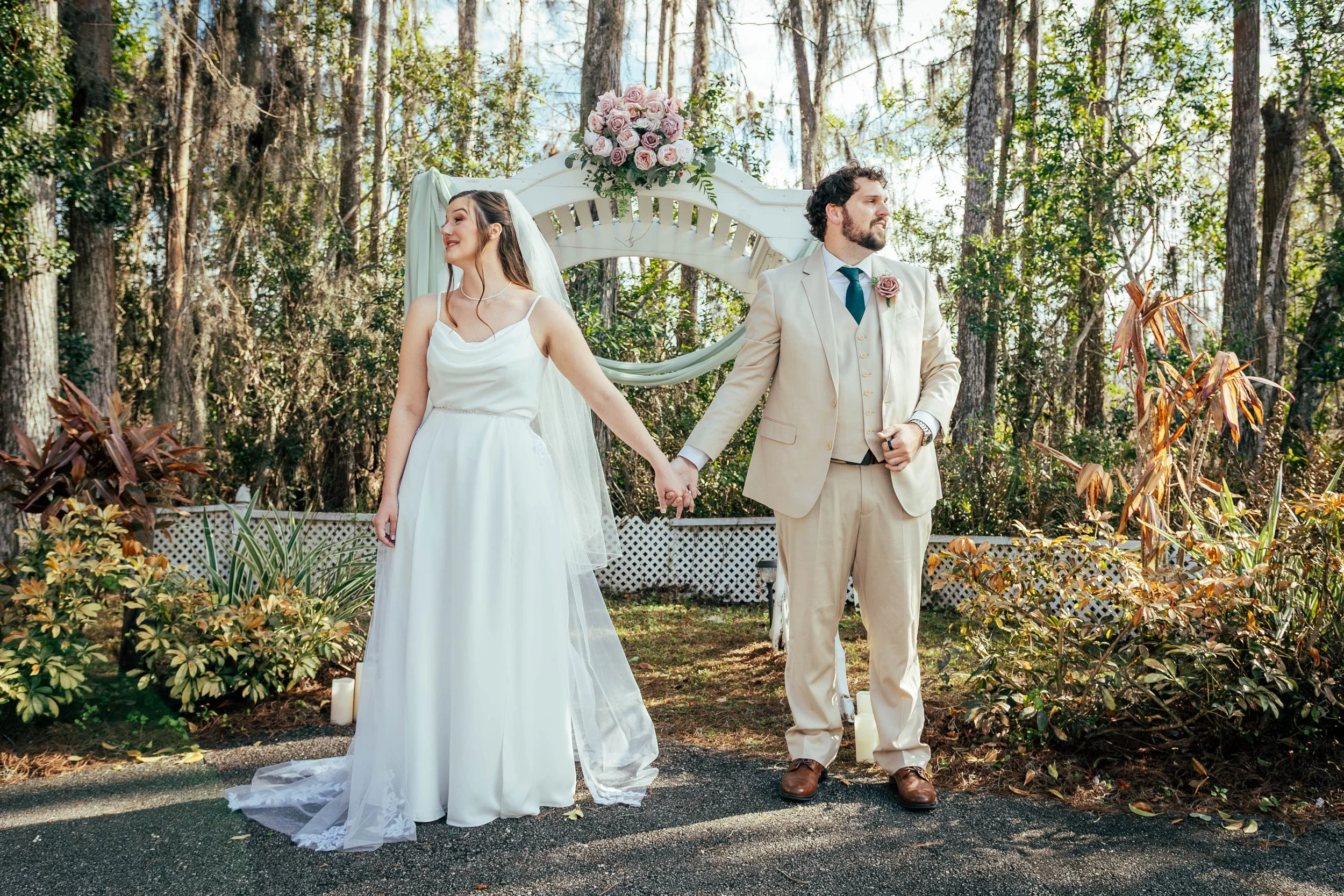 A bride and groom holding hands during an outdoor wedding ceremony in a wooded area, with a decorative floral arrangement and white lattice fence in the background.
