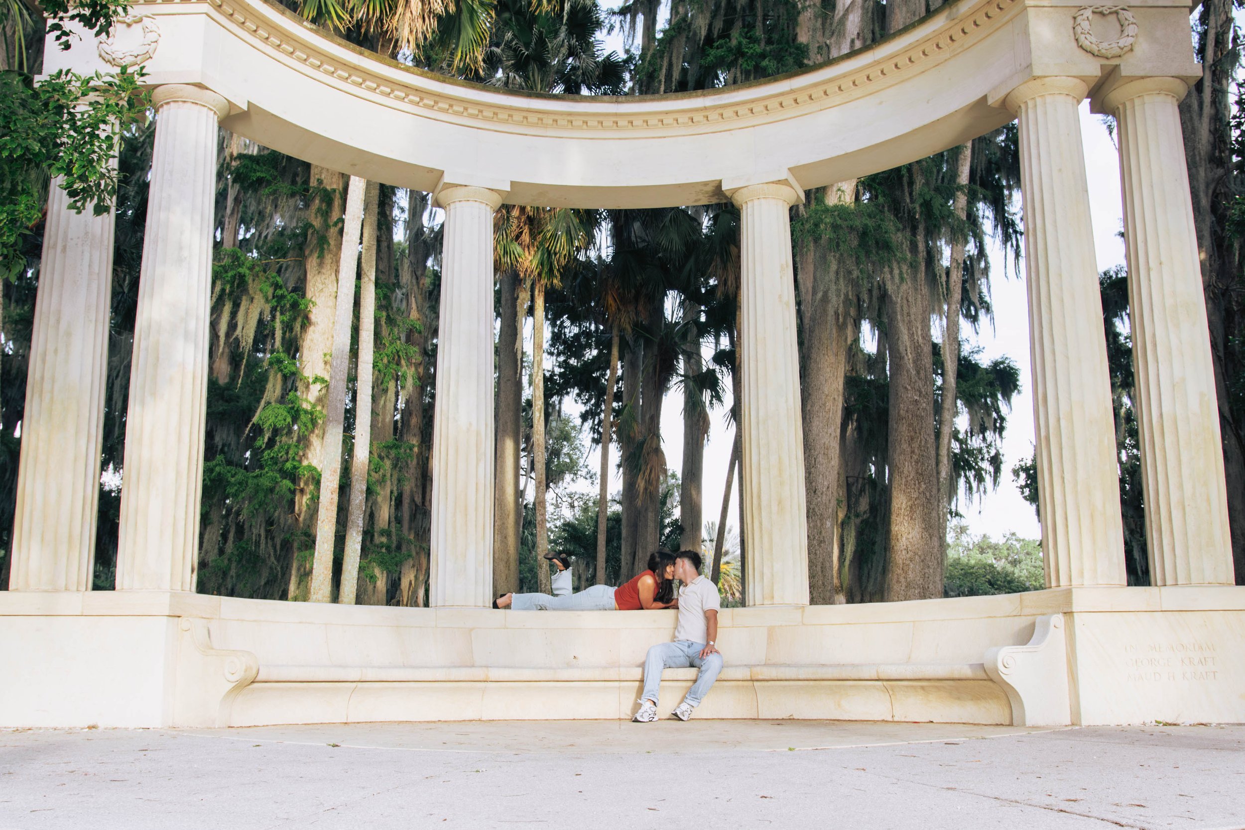 A couple kissing while lying on a marble ledge at a classical-style outdoor pavilion with white columns and a rounded roof, surrounded by tall trees.